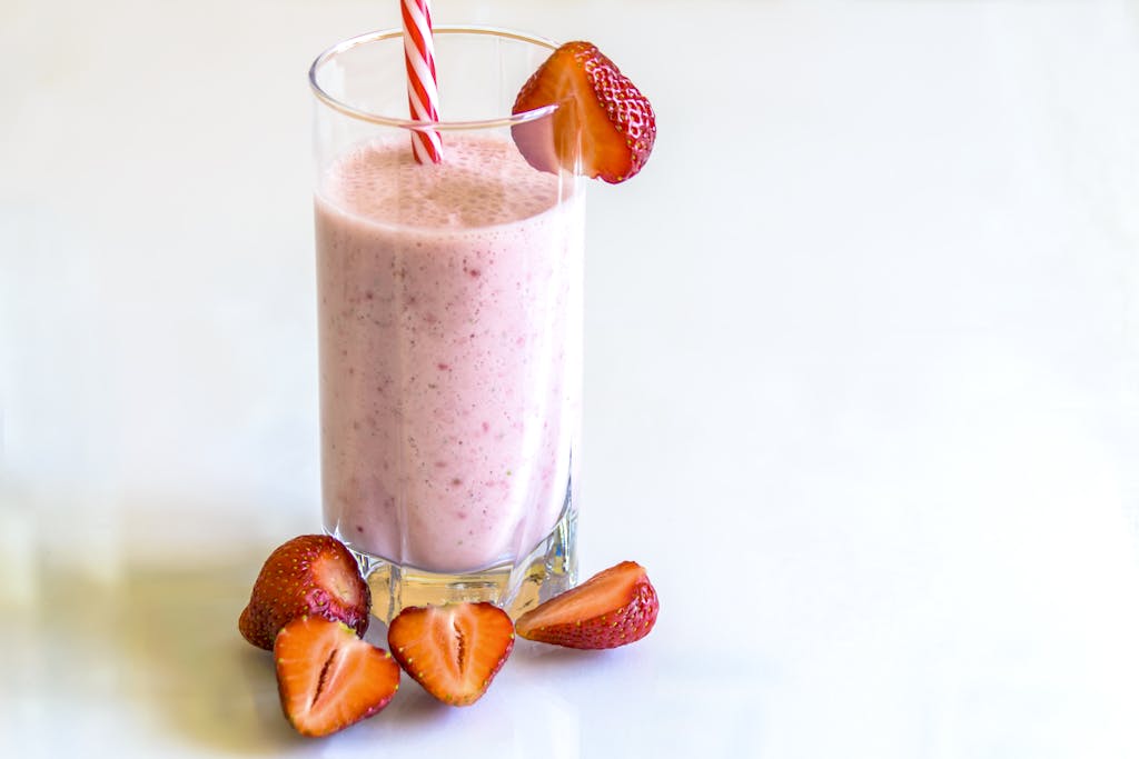 Delicious strawberry smoothie in a glass with fresh strawberries and a striped straw on a light background.