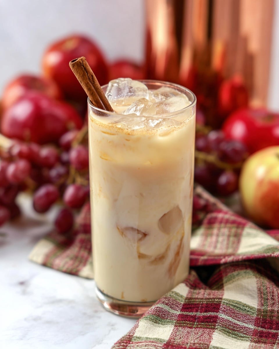 A tall clear glass filled with a creamy beige drink with visible ice cubes floating on top and a cinnamon stick placed inside at an angle. The drink has a smooth texture with some darker swirls near the bottom. The glass is set on a white marbled surface with a cloth underneath that has green, cream, and maroon plaid patterns. Behind the glass, there are red apples, red grapes, and a shiny copper container, all slightly out of focus. photo taken with an iphone --ar 4:5 --v 7