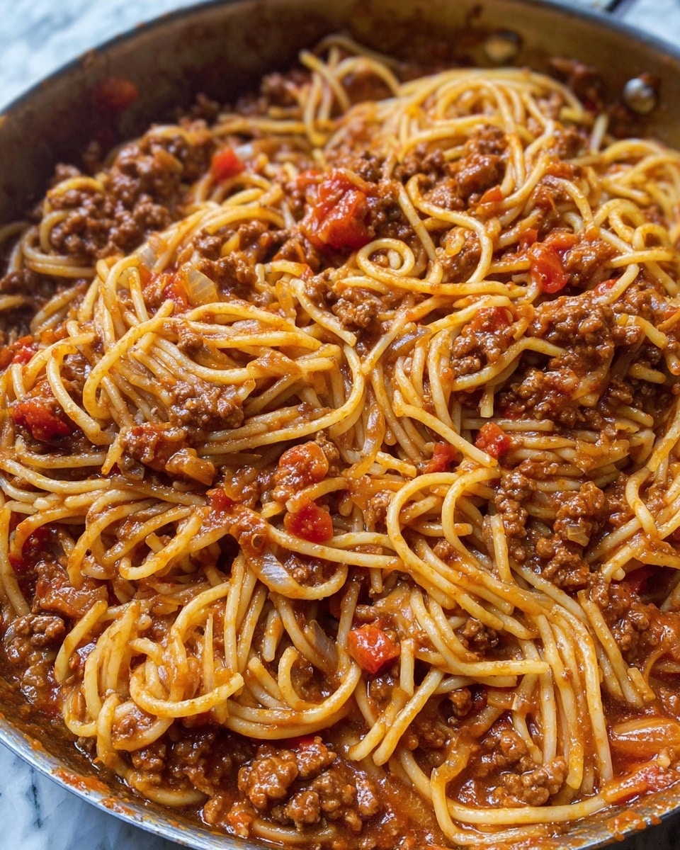 The image shows a close-up of spaghetti mixed with a thick meat sauce. The spaghetti noodles are long, light golden, and coated in a reddish-brown sauce made with ground meat and tomato chunks. The sauce contains visible pieces of cooked onion and small bits of red peppers, adding texture. The noodles and sauce are mixed evenly in a pan, with the sauce sitting slightly oily and glossy on the pasta. The background is a white marbled texture. photo taken with an iphone --ar 4:5 --v 7
