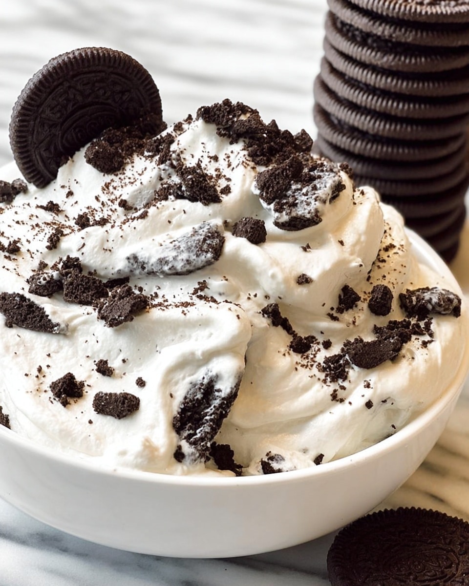 The dish is a creamy, thick white layer of whipped cream or mousse spread unevenly in a white bowl, topped with scattered black and dark brown cookie crumbs of varying sizes, some of which are large and broken pieces sitting on the surface. A large, dark brown and black cookie partially dipped in white cream stands upright on one side of the bowl. In the background, there is a tall stack of similar dark brown cookies on a white marbled surface. photo taken with an iphone --ar 4:5 --v 7