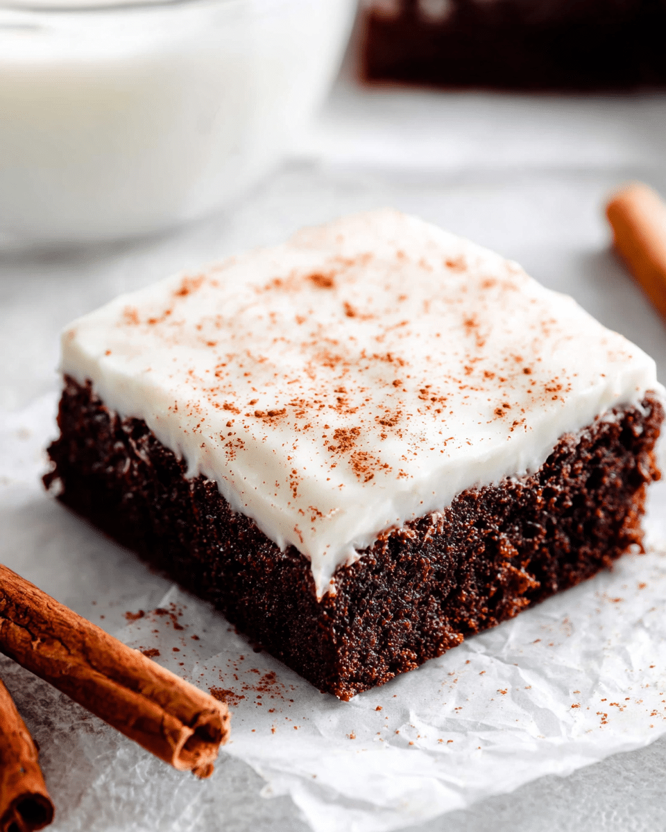 A single square piece of dark brown, moist chocolate cake forms the bottom layer, with a thick, smooth, white frosting layered evenly on top. The white frosting is sprinkled lightly with reddish-brown powder, likely cinnamon, scattered across its surface. The cake rests on white parchment paper, placed over a white marbled texture. There are two cinnamon sticks lying next to the cake in the bottom left corner. In the blurred background, a glass bowl with white contents is partly visible at the top left. photo taken with an iphone --ar 4:5 --v 7
