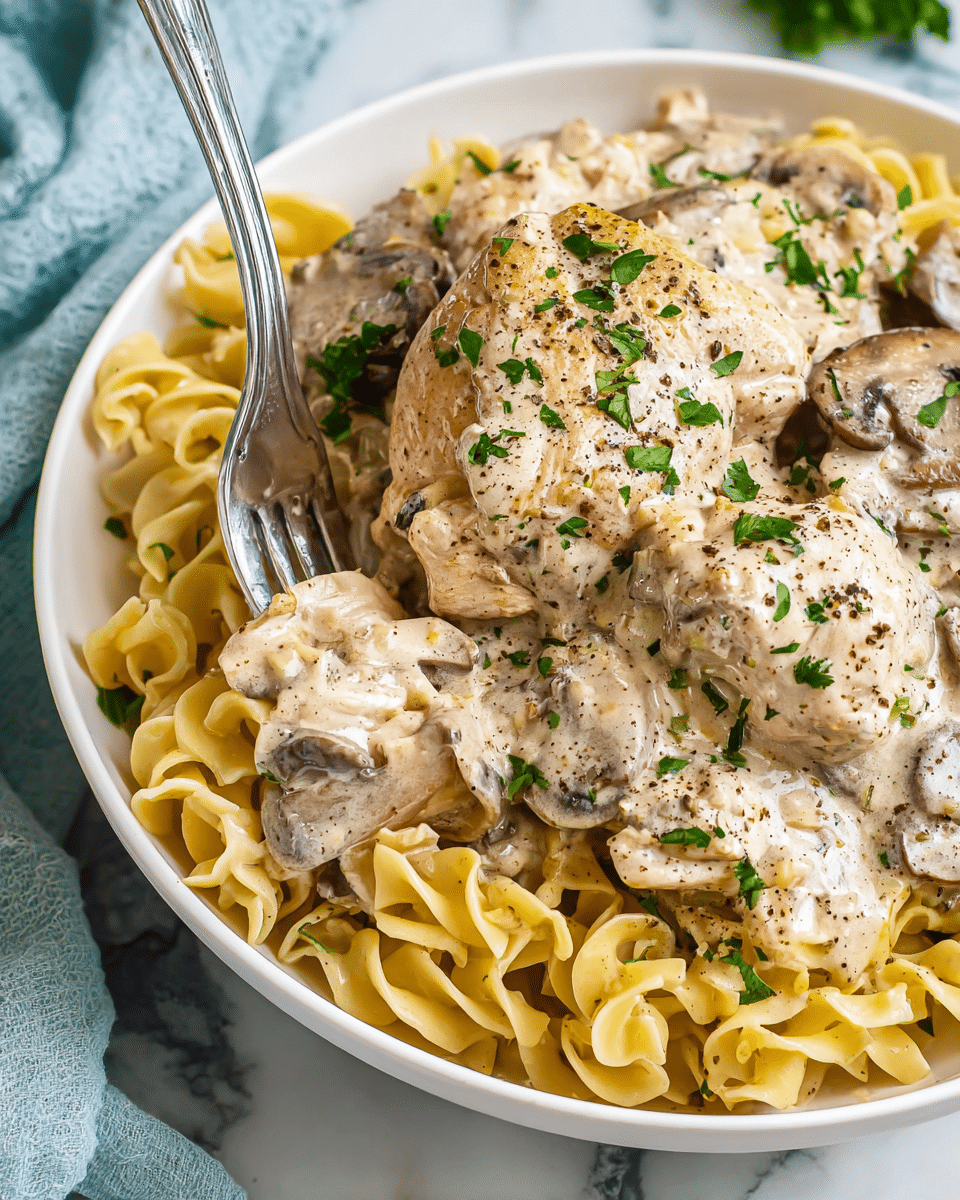 A white bowl filled with a creamy chicken and mushroom pasta dish, showing two main layers: at the bottom, golden-yellow curly pasta noodles with a soft texture, and on top, beige chicken pieces covered in a thick creamy mushroom sauce with sliced brown mushrooms scattered throughout, sprinkled with fresh green parsley and black pepper for contrast, with a silver fork partly inserted into the pasta, the scene set on a white marbled surface with a light blue cloth partially visible. Photo taken with an iphone --ar 4:5 --v 7
