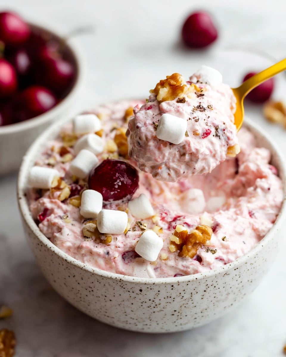 A close-up view of a white speckled bowl filled with a creamy, light pink textured dessert that has a thick, fluffy appearance. The dessert contains small red cherries and crunchy pieces of walnut scattered on top and mixed inside. White mini marshmallows are also placed both on the surface and stirred into the dessert. A yellow spoon lifting a large scoop shows the dense mix with visible cherries, walnuts, and marshmallows. In the background, there is a white bowl with additional whole cherries slightly out of focus, all set on a white marbled surface. Photo taken with an iphone --ar 4:5 --v 7