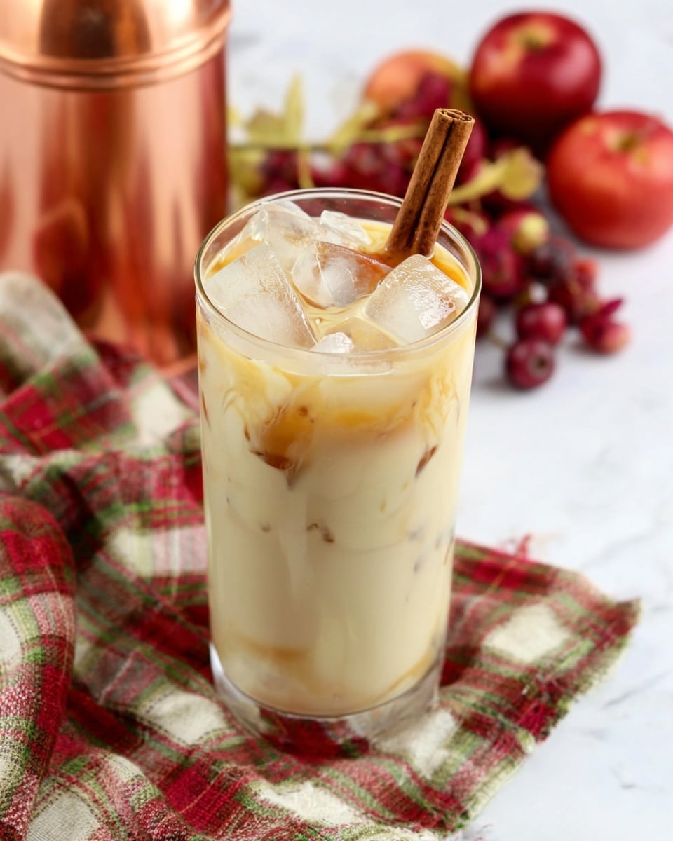 A tall clear glass filled with a creamy, light beige drink showing visible brown swirls near the bottom, topped with several large ice cubes floating inside and a cinnamon stick resting diagonally on top. The glass sits on a crumpled plaid fabric with green, red, cream, and brown colors, all placed on a white marbled surface. In the background, several red apples and some small red fruits add a pop of color, along with a shiny copper container on the left side. photo taken with an iphone --ar 4:5 --v 7