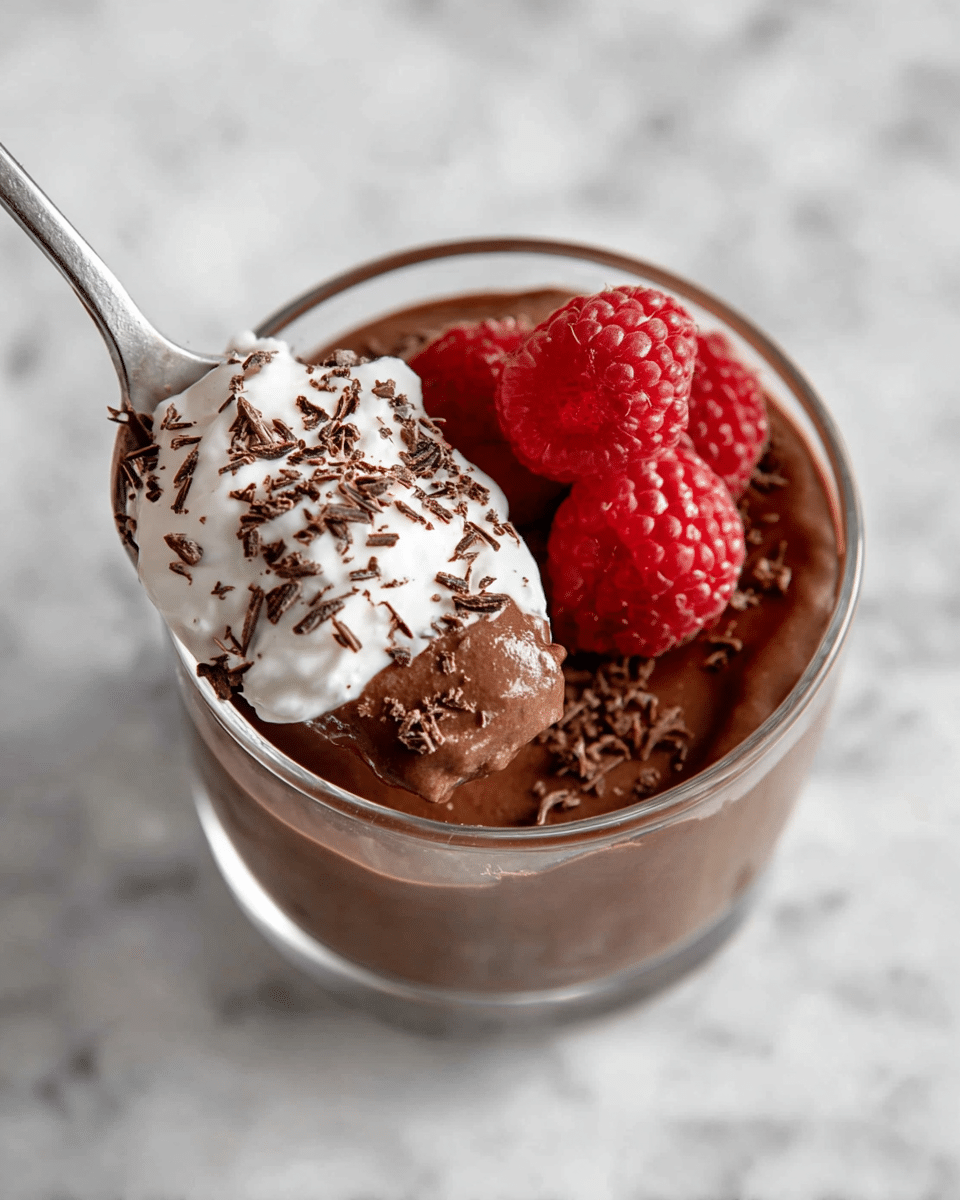 A clear glass cup contains a rich, smooth dark brown chocolate mousse as the main layer. On top, there is a dollop of white whipped cream placed off-center to one side. Two fresh, bright red raspberries sit beside the whipped cream, adding color contrast and texture. A silver spoon is dipped into the mousse, holding a scoop that shows a mix of creamy chocolate and white whipped cream sprinkled with fine dark chocolate shavings. The cup is set on a white marbled surface, enhancing the visual appeal. photo taken with an iphone --ar 4:5 --v 7