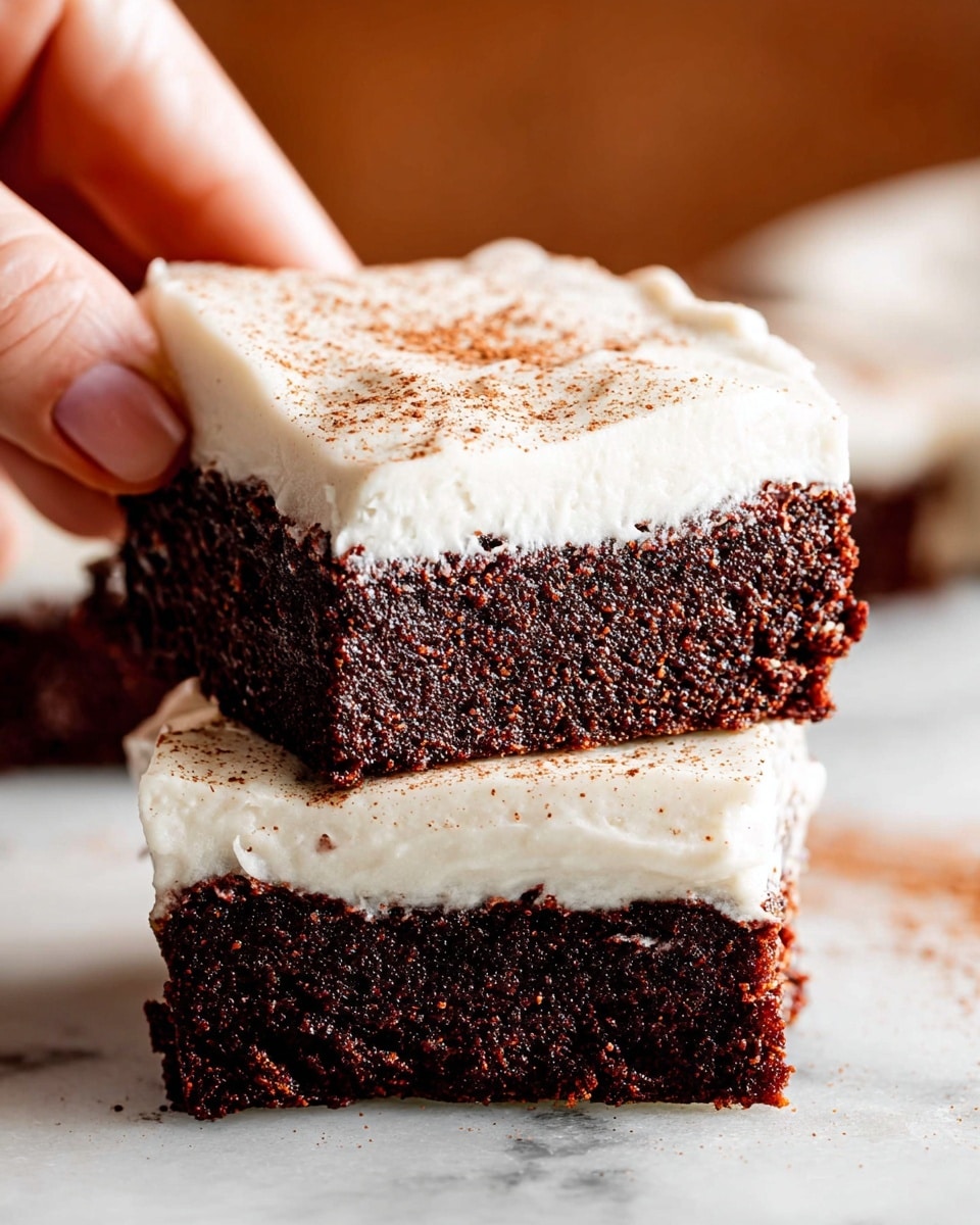 The image shows two thick square brownies stacked on top of each other on a white marbled surface. Each brownie has two clear layers: a dark, rich chocolate base with a dense, crumbly texture, and a thick, smooth layer of white frosting on top, which is sprinkled lightly with brown powder, likely cinnamon or cocoa. A woman's hand is gently holding the top brownie from the side with fingers visible. The background is softly blurred with warm brown tones. Photo taken with an iphone --ar 4:5 --v 7
