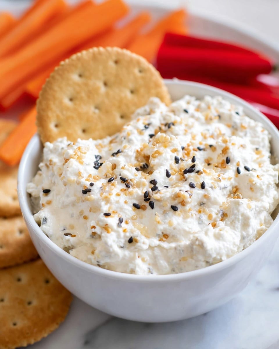 A white bowl filled with a creamy, thick dip that has a lumpy texture, looking like cottage cheese mixed with herbs and spices. The dip is topped with small black seeds and tiny golden brown bits, evenly spread over the surface. A round, light brown cracker is placed partially inside the dip on the left side. In the background, there are blurred baby carrots and red pepper sticks on a white marbled surface. photo taken with an iphone --ar 4:5 --v 7