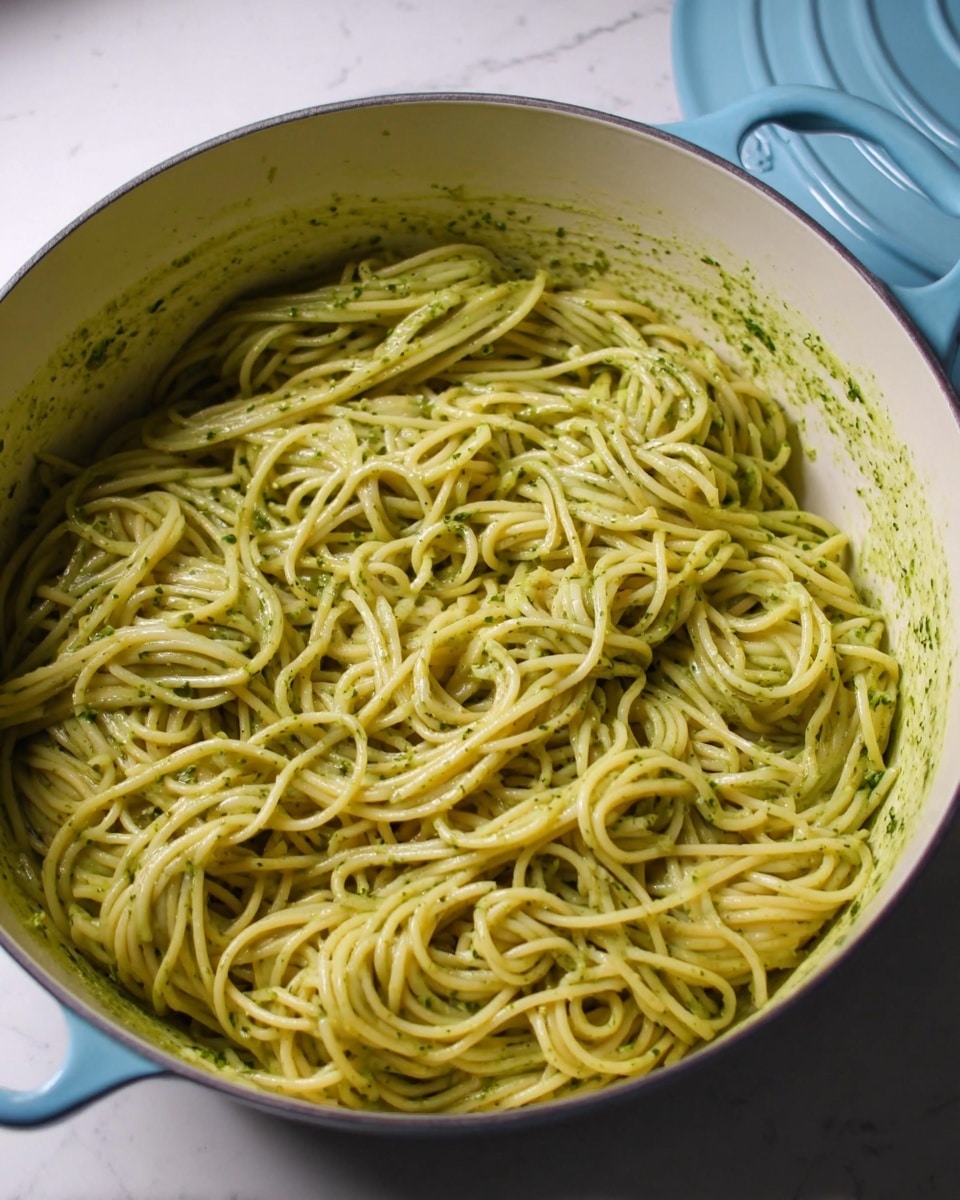 A large white pot filled with spaghetti pasta coated in a smooth, light green sauce that clings to each strand. The spaghetti is twisted and piled loosely, showing the sauce’s creamy texture with small bits of green herbs throughout. The edges of the pot show some sauce splattered, emphasizing the fresh mix. The pot lid, colored pale blue, rests nearby on a white marbled surface. photo taken with an iphone --ar 4:5 --v 7