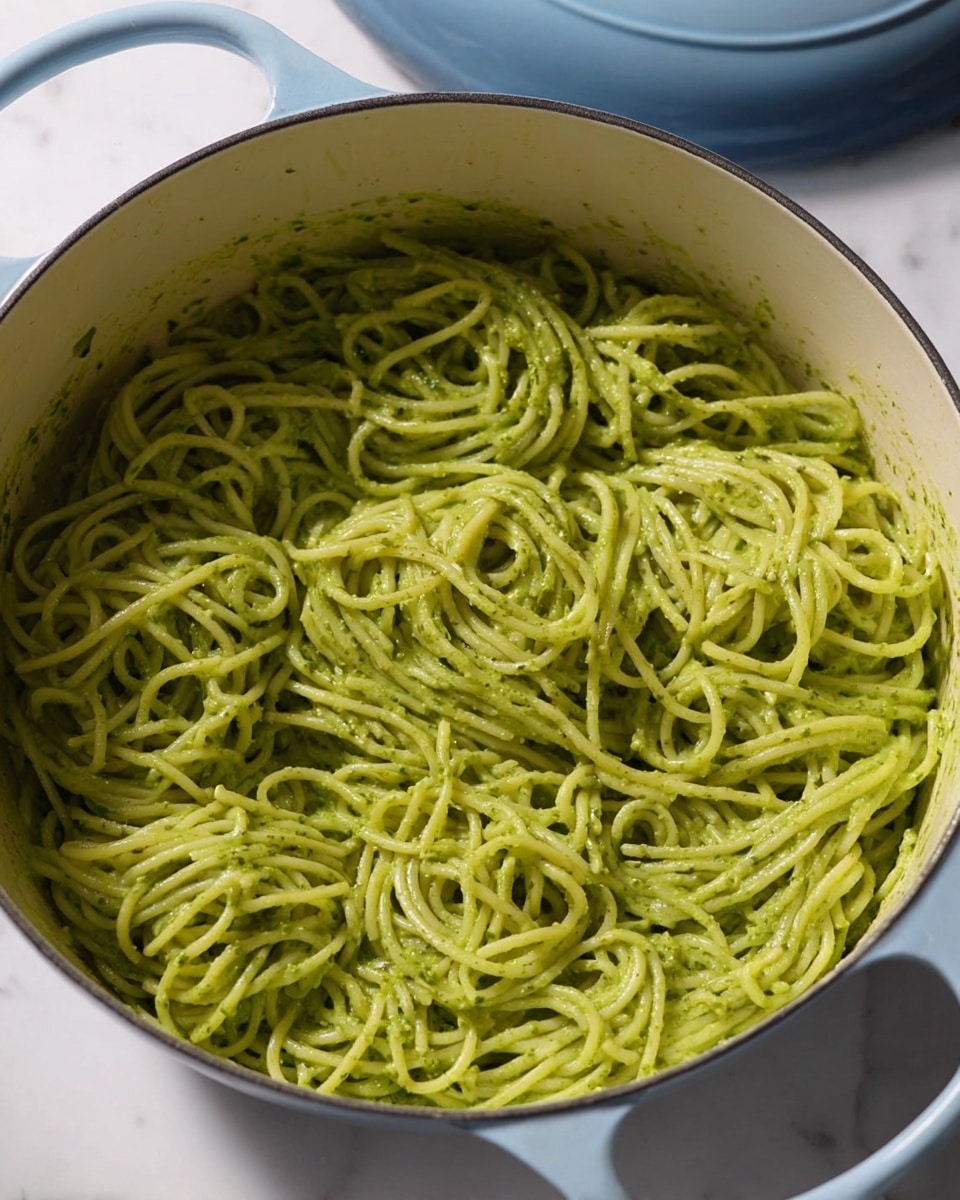 A close-up view of spaghetti noodles mixed thoroughly with a smooth, creamy green sauce, evenly coating each strand. The noodles are tangled and layered in a round white cast iron pot with light blue handles and a lid partially visible in the background. The sauce has a vibrant green color with a slightly textured surface, indicating a fresh herb or vegetable blend. The pot sits on a clean white marbled surface, highlighting the bright green pasta inside. photo taken with an iphone --ar 4:5 --v 7