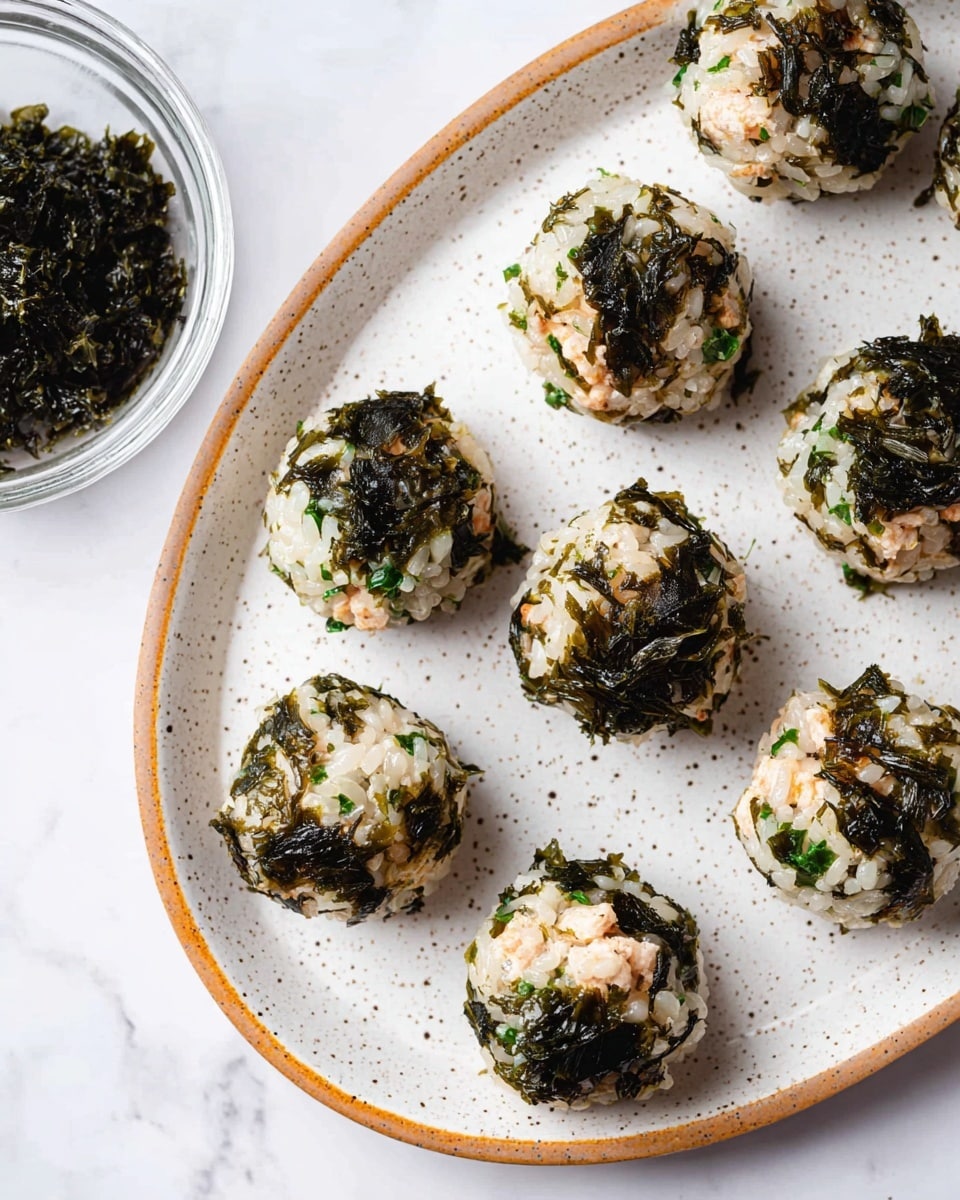 Nine round rice balls covered with chopped green seaweed sit arranged on a white speckled plate with a thin orange-brown rim, all placed on a white marbled surface. Each ball has a base layer of white rice mixed with small chunks of light brown filling, then coated unevenly with a layer of dark green, leafy seaweed that adds texture and color contrast. In the top left corner, there is a small clear glass bowl filled with more dark green seaweed, partially visible. photo taken with an iphone --ar 4:5 --v 7