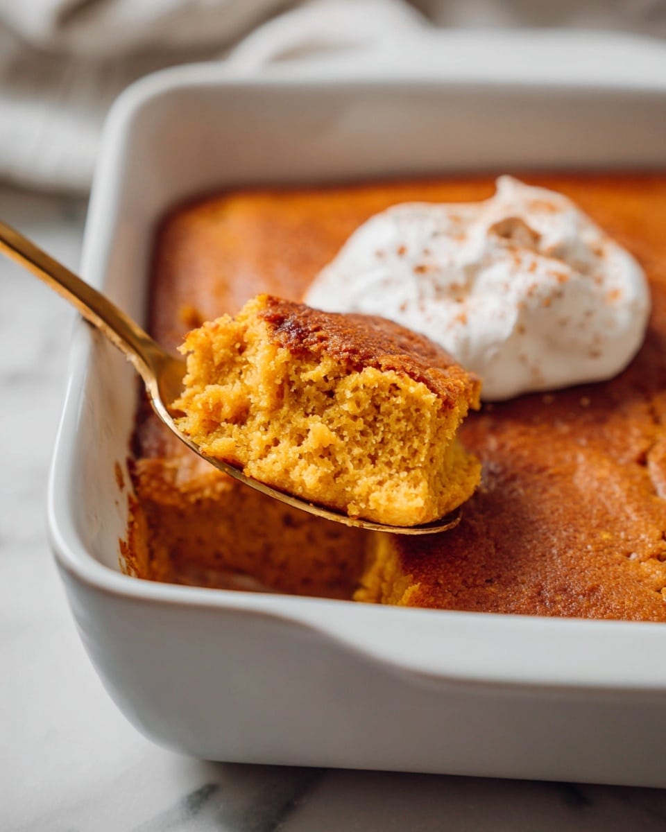 A close-up of a white square baking dish filled with a single-layer, golden brown soft pumpkin dessert with a sponge-like texture. A golden spoon lifts a piece from the dish, showing the crumbly, moist inside of the dessert. On one corner of the dessert, there is a dollop of white whipped cream with a sprinkle of light brown powder on top. The dish rests on a white marbled surface. photo taken with an iphone --ar 4:5 --v 7