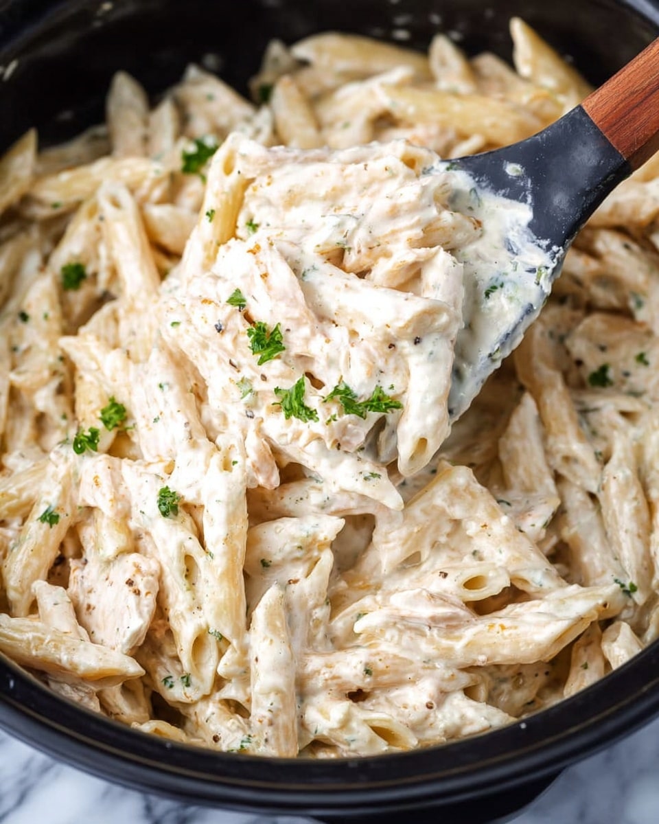 The image shows a close-up of creamy pasta mixed in a black pot. The dish has one main layer of penne pasta coated in a thick, white creamy sauce that looks smooth and rich, with small bits of herbs and spices visible in the sauce. Light green chopped parsley pieces are scattered on top of the pasta, adding a small touch of color. A black spoon with a wooden handle is lifting a portion of the pasta, showing the texture of the sauce and the penne clearly. The background surface is a white marbled texture. photo taken with an iphone --ar 4:5 --v 7