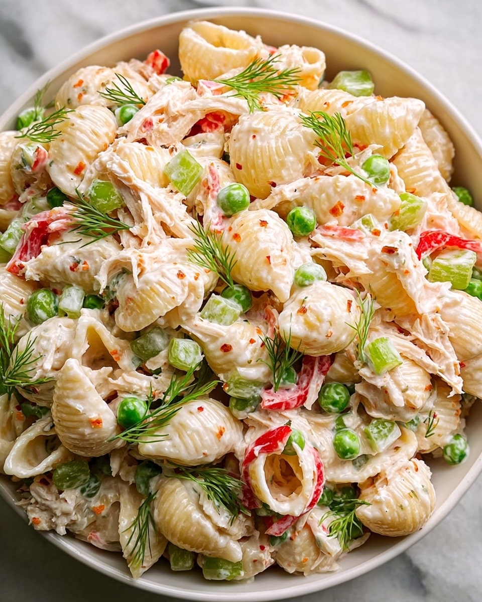The dish is a creamy pasta salad served in a white bowl, placed on a white marbled surface. It has large shell pasta coated in a white creamy dressing, mixed with small green peas, light green celery chunks, red bell pepper strips, and shredded white chicken. Small sprigs of fresh green dill are scattered throughout, adding a vibrant contrast. The textures range from smooth and creamy to crunchy and fresh, with the pasta shells curling to hold the mix. Photo taken with an iphone --ar 4:5 --v 7