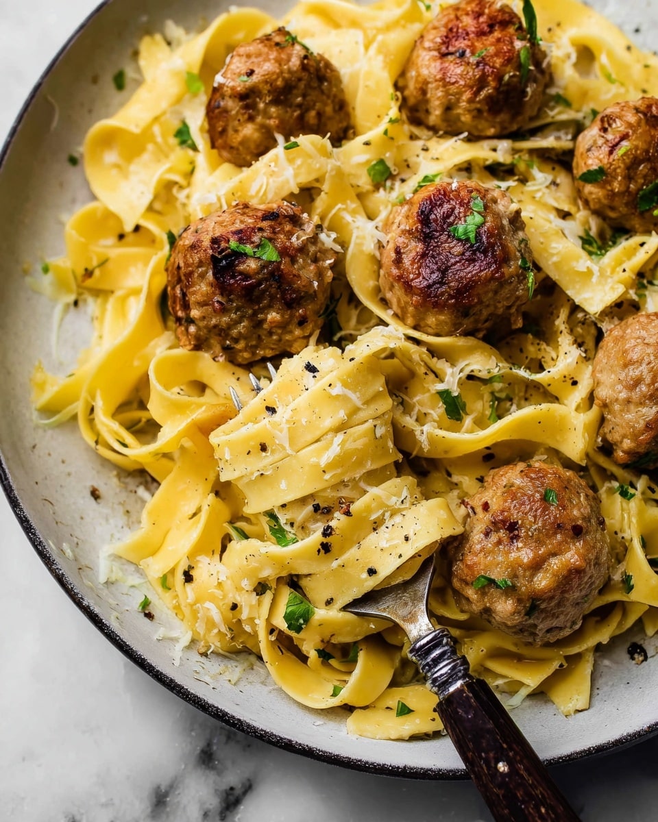 The image shows a close-up of pasta and meatballs served on a white plate. There are several large, golden-brown meatballs, with a textured and slightly crispy outside, scattered among thick, flat yellow pasta noodles. The pasta is coated lightly with what looks like a creamy sauce and sprinkled with small green herb pieces. There are also black pepper flakes visible on the noodles and meatballs. A fork with a dark wooden handle lifts some pasta from the plate, adding a sense of movement to the dish. The plate rests on a white marbled surface. photo taken with an iphone --ar 4:5 --v 7