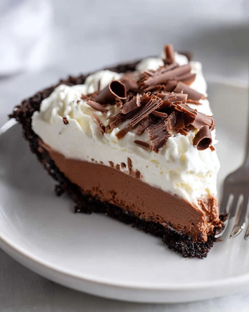 A slice of three-layer pie is shown on a white plate resting on a white marbled textured surface. The bottom layer is a dark, crumbly crust, almost black in color. Above that is a thick, smooth layer of rich brown chocolate filling. On top is a fluffy and airy white cream layer, crowned with several curled dark chocolate shavings scattered evenly. A silver fork is placed behind the pie slice on the plate. photo taken with an iphone --ar 4:5 --v 7