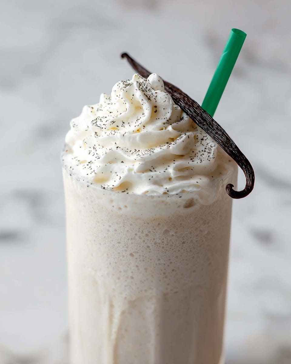 A tall clear glass filled with a creamy light beige milkshake topped with a thick layer of white whipped cream, sprinkled with tiny black vanilla seeds, and decorated with a dark brown vanilla pod curved on top. A green straw is placed into the milkshake on one side. The drink has foam dripping slightly down the edge of the glass. The glass is set against a white marbled texture background. Photo taken with an iphone --ar 4:5 --v 7
