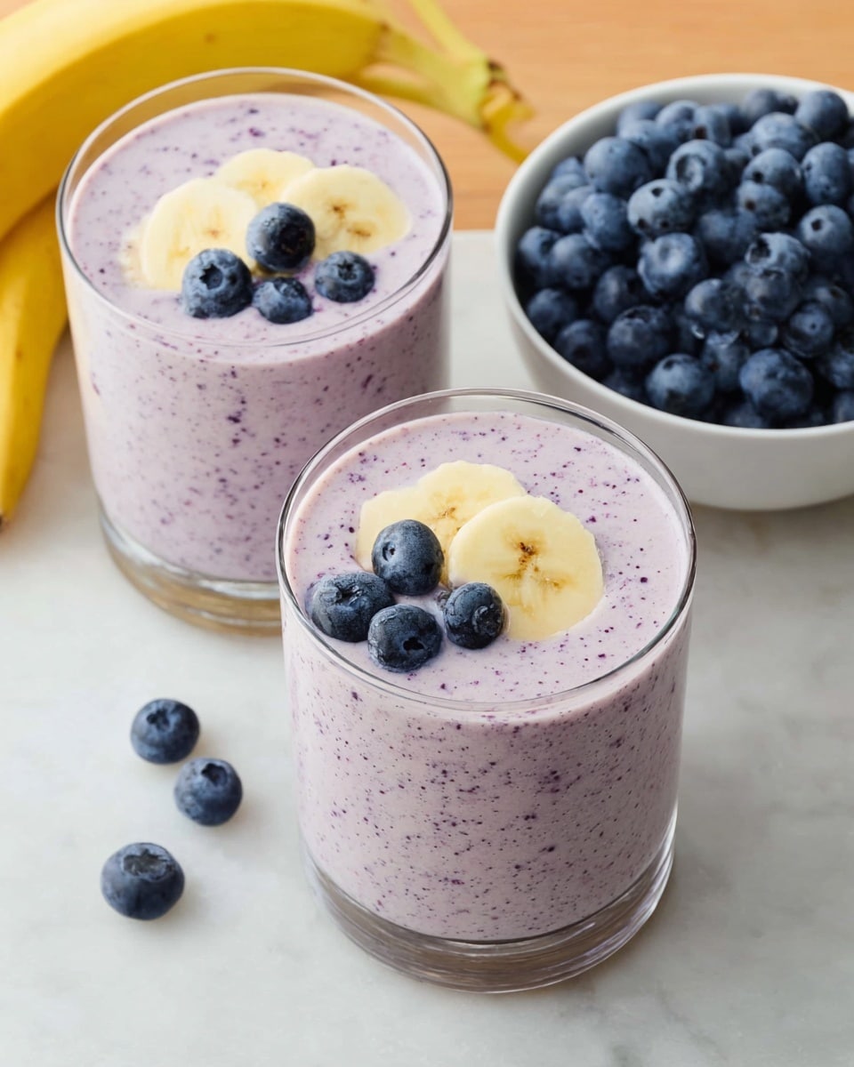 The image shows a glass filled with a thick, light purple smoothie speckled with tiny dark purple bits, topped with two whole blueberries and two thin round slices of banana placed near the glass edge. Behind it, there is a white bowl full of fresh blueberries, and another glass with the same smoothie and a single blueberry floating on top. The setting is on a light wood surface with a few scattered blueberries around. The background features a yellow banana slightly out of focus. photo taken with an iphone --ar 4:5 --v 7