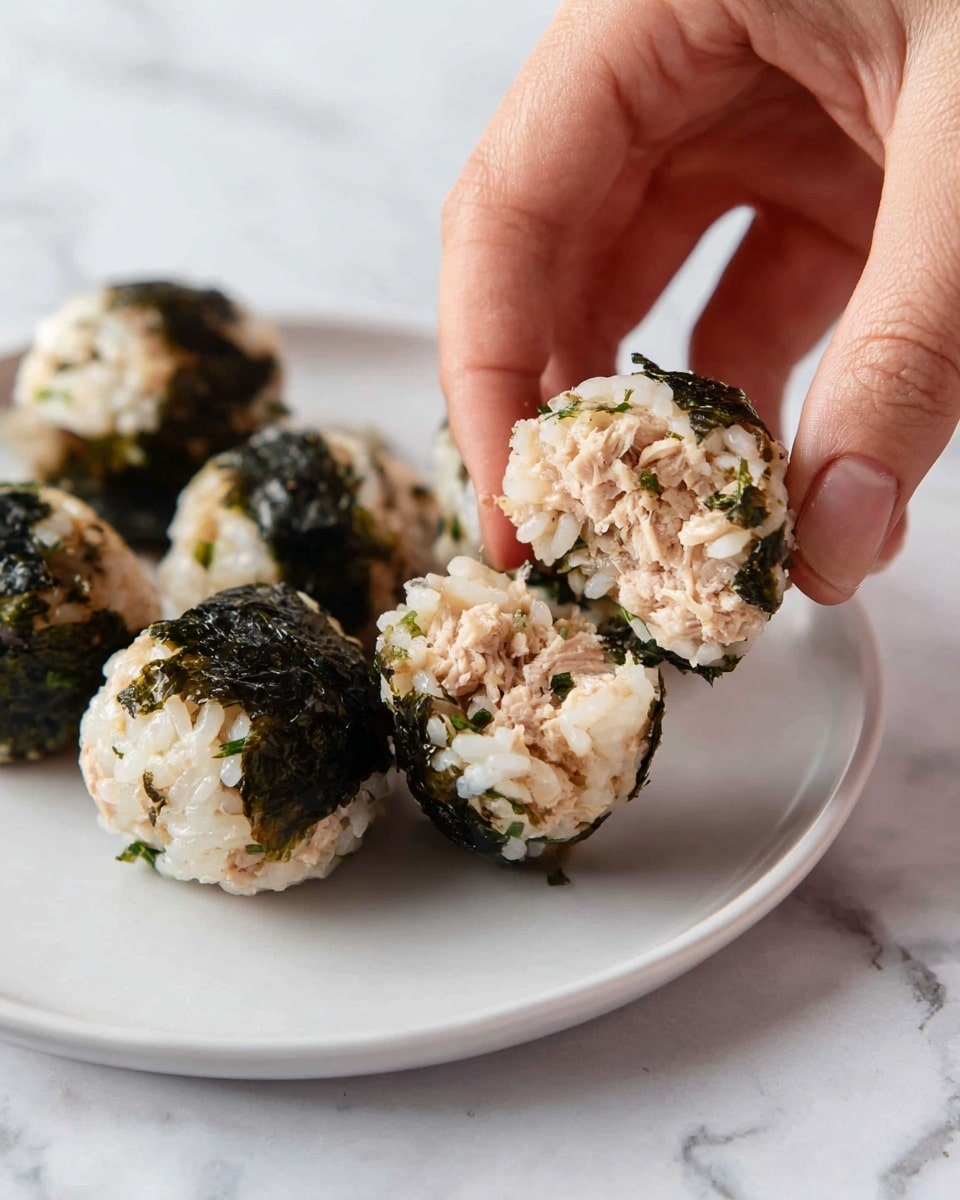 A close-up view shows a white plate on a white marbled surface, holding several small rice balls covered in dark green seaweed flakes. One rice ball is being split in two by a woman's hand, revealing the inside layers of cooked white rice mixed with light brown tuna chunks, with some small green herb bits. The texture is slightly sticky and moist, with the outer layer of seaweed giving a rough dark green outline. Photo taken with an iphone --ar 4:5 --v 7