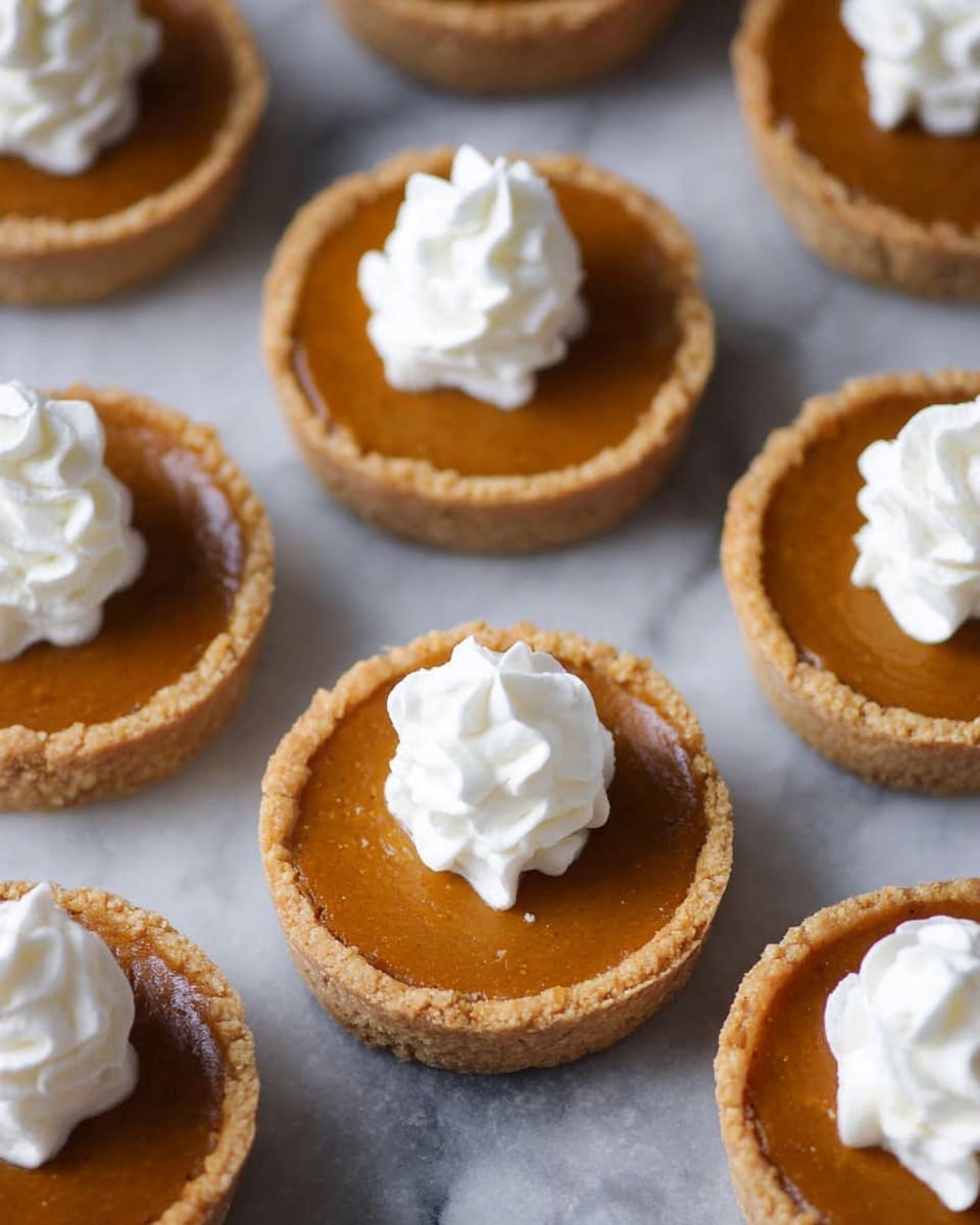 The image shows several small round pumpkin pies with two visible layers: a light brown crust base and a smooth, darker orange pumpkin filling on top. Each pie has a small, fluffy dollop of white whipped cream placed in the center. The pies are closely arranged on a white marbled textured surface. Photo taken with an iphone --ar 4:5 --v 7