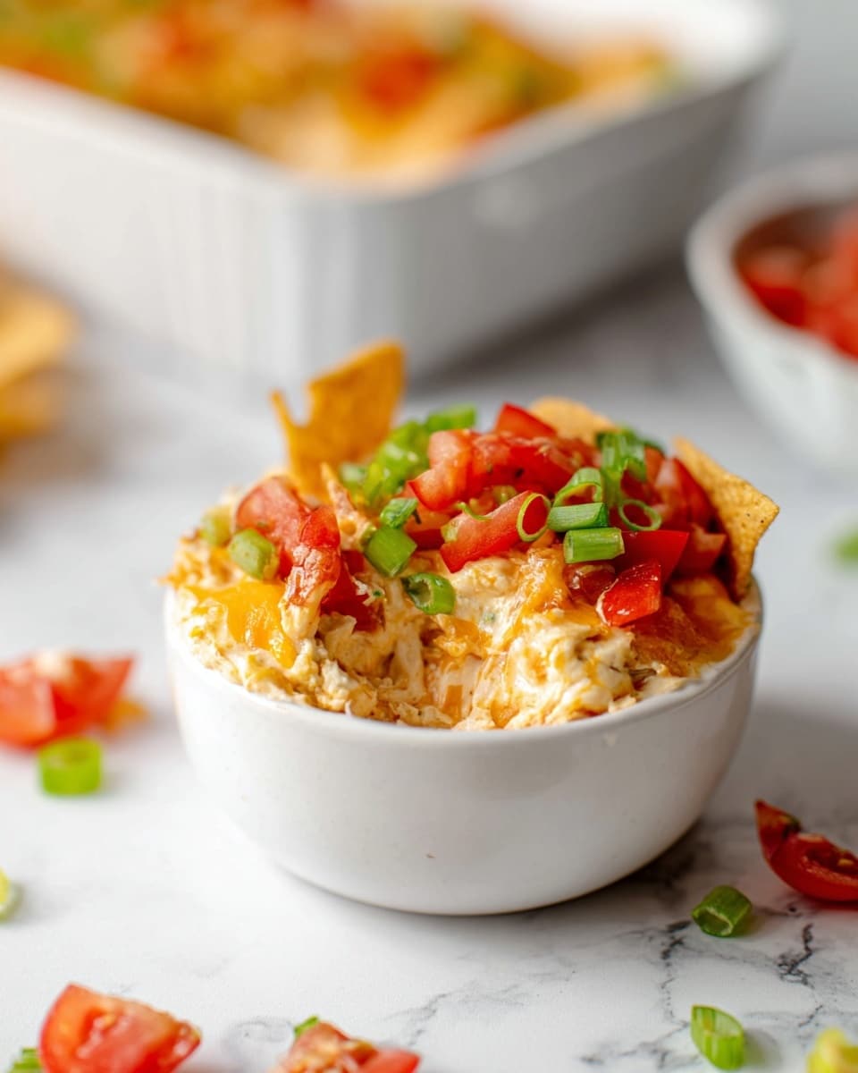 A white bowl filled with a creamy layered dip topped with golden crunchy chips, bright red diced tomatoes, and fresh green sliced scallions. The dip appears thick and cheesy with a smooth texture beneath the chips. The bowl is placed on a white marbled surface with scattered diced tomatoes and scallions around it. The background shows a blurred white baking dish with more of the same dip inside. Photo taken with an iphone --ar 4:5 --v 7