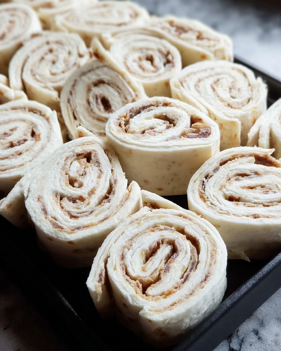 A close-up view of multiple small pinwheel sandwiches made from soft white flatbread rolled with a thin layer of light brown filling, likely meat or spread, forming visible spiral layers. Each sandwich shows about four to five white and brown swirled layers tightly rolled and sliced into uniform bite-sized pieces. The pinwheels are arranged closely on a flat black rectangular tray placed on a white marbled surface, and the texture of the flatbread looks soft and smooth with slight irregularities. photo taken with an iphone --ar 4:5 --v 7