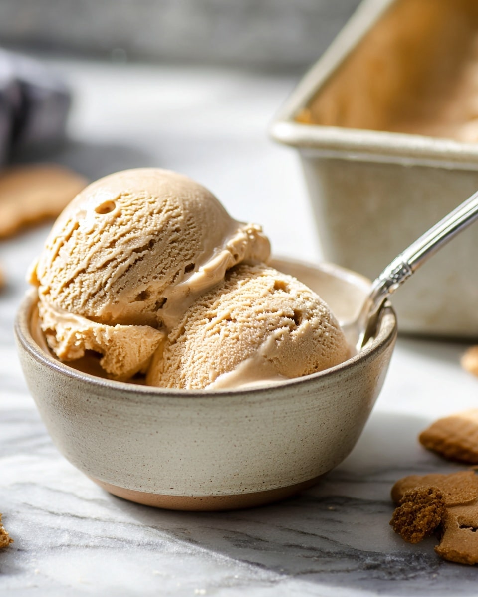 The image shows two scoops of smooth, creamy light brown ice cream in a small, round beige ceramic bowl. The scoops have a slightly rough texture with some small air holes and light melting edges. The bowl sits on a white marbled surface with some shadows, and there is a silver spoon inside the bowl. In the blurred background, there is a rectangular beige container and some scattered cookie pieces on the right side, adding to the cozy feel. photo taken with an iphone --ar 4:5 --v 7