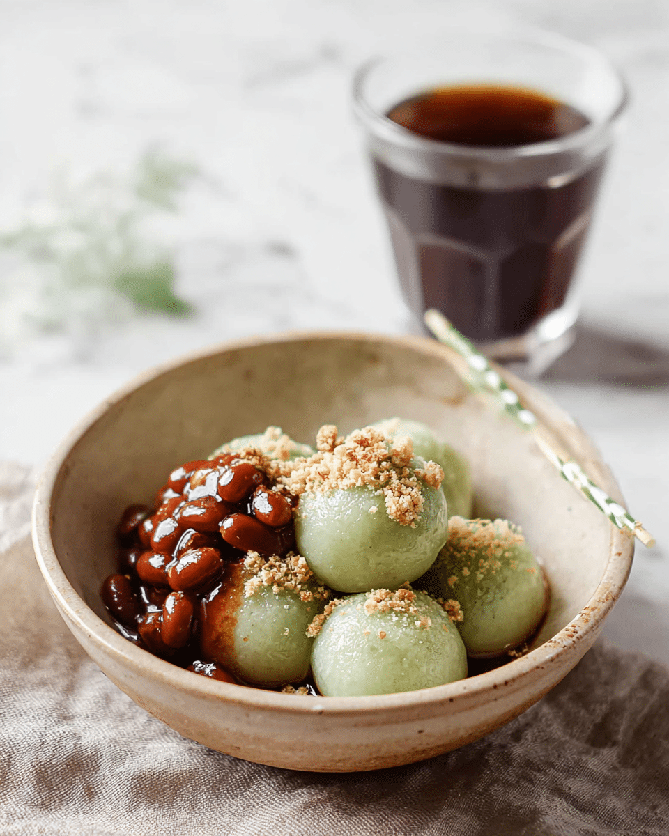 The image shows a rustic beige bowl placed on a white marbled textured surface, filled with six round, pale green mochi balls stacked close to each other. On the left side of the mochi, there is a glossy reddish-brown topping of cooked beans that looks sticky and shiny. A sprinkle of light beige crushed nuts or powder is scattered over the mochi and beans, adding texture and contrast. Resting in the bowl is a thin stick with green and white stripes, partially inserted among the mochi. In the background, there is a transparent glass filled halfway with dark brown coffee, slightly out of focus. photo taken with an iphone --ar 4:5 --v 7