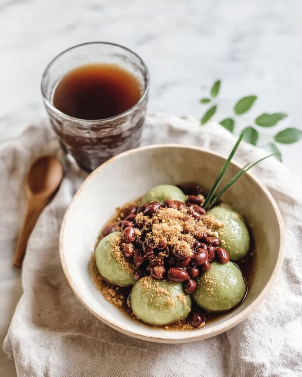 The image shows a white bowl filled with four round, green-colored dumplings placed closely together in the center. On top of the dumplings, there is a generous layer of small red beans that look soft and sticky, and a sprinkling of light tan powder or crumbs adds texture over the beans and dumplings. A thin green leaf or grass is placed on the right side of the dumplings inside the bowl. Next to the bowl, there is a clear glass cup filled with a dark brown liquid, likely a tea or coffee, set on a white marbled surface with a light cloth underneath the bowl. The overall colors are soft and natural, with a cozy and simple feel. photo taken with an iphone --ar 4:5 --v 7