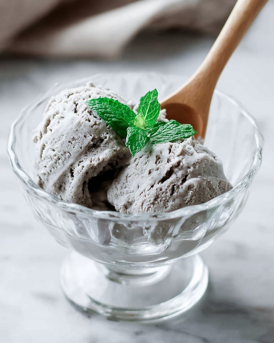 Two scoops of light gray ice cream with a rough, creamy texture sit in a clear glass bowl, with a small bright green mint leaf on top for decoration. A light wooden spoon is placed inside the bowl behind the ice cream. This bowl rests on a white marbled surface. photo taken with an iphone --ar 4:5 --v 7