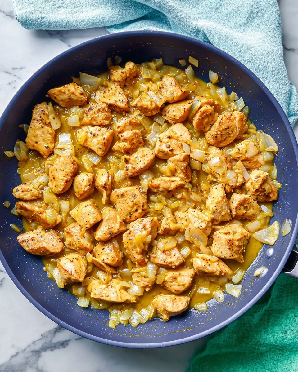 The image shows a dark blue frying pan filled with cooked chicken pieces and chopped onions in a thick golden-yellow sauce. The chicken pieces are lightly browned and scattered evenly throughout the pan, mixed with translucent diced onions. The sauce surrounds and partly covers the chicken and onions, giving a slightly glossy texture. The pan is placed on a white marbled surface with a folded light blue cloth in the upper part of the image and a green cloth partially visible in the lower right corner. photo taken with an iphone --ar 4:5 --v 7