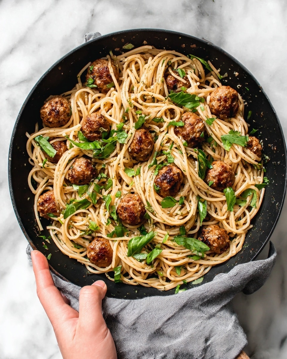 A black skillet filled with a mixture of light brown spaghetti noodles and round, golden-brown meatballs evenly spread throughout. Bright green leafy herbs are scattered over the pasta and meatballs, adding a fresh contrast. The skillet is held by a woman's hand covered with a gray cloth at the handle, resting on a white marbled surface. The dish looks hearty and fresh with a mix of soft pasta strands and crispy, browned meatballs. photo taken with an iphone --ar 4:5 --v 7