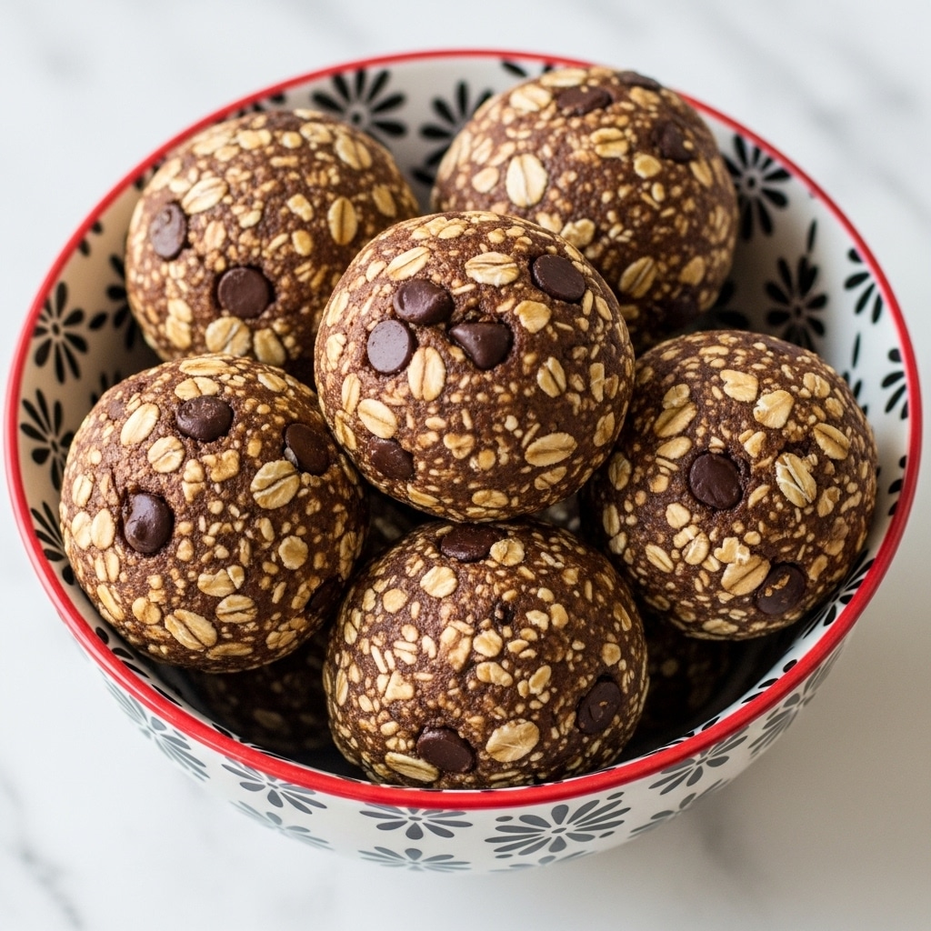 Several round, brown oatmeal balls with visible oats and dark chocolate chips are piled inside a white bowl with black floral patterns and a red rim. The texture of the balls looks rough and crumbly, with layers of oats and chocolate chips mixed in evenly. The bowl is placed on a white marbled surface. Photo taken with an iphone --ar 4:5 --v 7