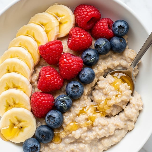 A close-up view of a bowl of creamy oatmeal with a slightly lumpy texture forming the bottom layer, topped with a row of thin, pale yellow banana slices on the left side, a cluster of bright red raspberries in the middle, and several small, round, dark blue blueberries scattered around the raspberries and oatmeal. Golden honey is drizzled lightly over the oatmeal and fruits, glistening under the light. The bowl is white, and a silver spoon rests inside it on the right side, partially scooping some oatmeal. The background is a white marbled texture. photo taken with an iphone --ar 4:5 --v 7