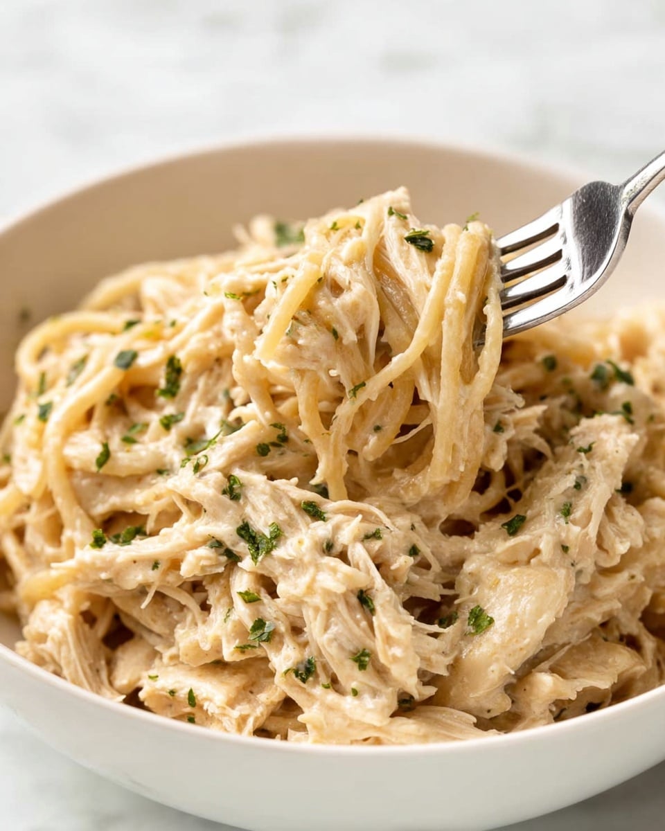 A close-up view of a white bowl filled with creamy, shredded chicken pasta that has a pale beige color mixed with small green herb flakes scattered on the top and throughout the dish. The soft pasta strands and tender chicken pieces are coated in a smooth, thick sauce. A silver fork is inserted into the pasta from the top right, with its tines lifting some of the noodles. The bowl sits on a white marbled surface, providing a clean and bright background. photo taken with an iphone --ar 4:5 --v 7