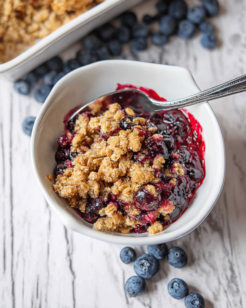 In a white bowl, there is a layered dessert consisting of a deep purple-blue blueberry filling at the bottom, topped with a golden-brown crunchy oat crumble scattered unevenly over the fruit layer. The crumble has varying sizes of clumps, giving a textured contrast to the smooth, glossy fruit layer beneath. A silver spoon is placed in the bowl, partly covered by the crumble topping. Fresh blueberries are scattered around the bowl on a white marbled surface, adding more color to the scene. Photo taken with an iphone --ar 4:5 --v 7