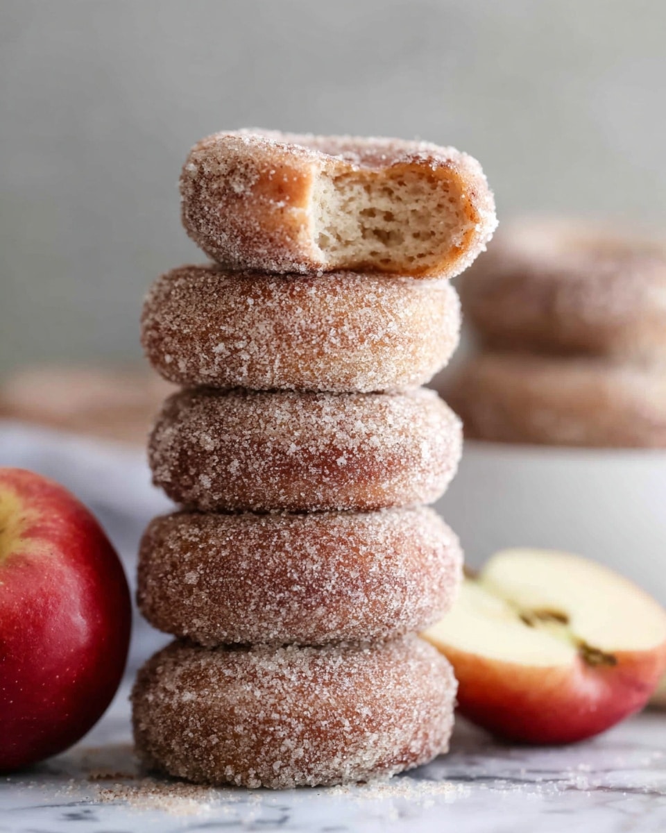 A stack of five round donuts coated in a light layer of granulated sugar stands tall on a white marbled surface, with the top donut having a bite taken out, revealing a soft, grainy beige inside. Next to the stack, there is a smaller donut hole covered in the same sugar coating and a whole, red apple with a smooth, shiny surface, accompanied by a half apple showing its pale juicy inside. In the blurred background, a white bowl and another sugar-coated donut are visible. The overall setting is simple and clean, highlighting the textures of the sugar and fruit. photo taken with an iphone --ar 4:5 --v 7