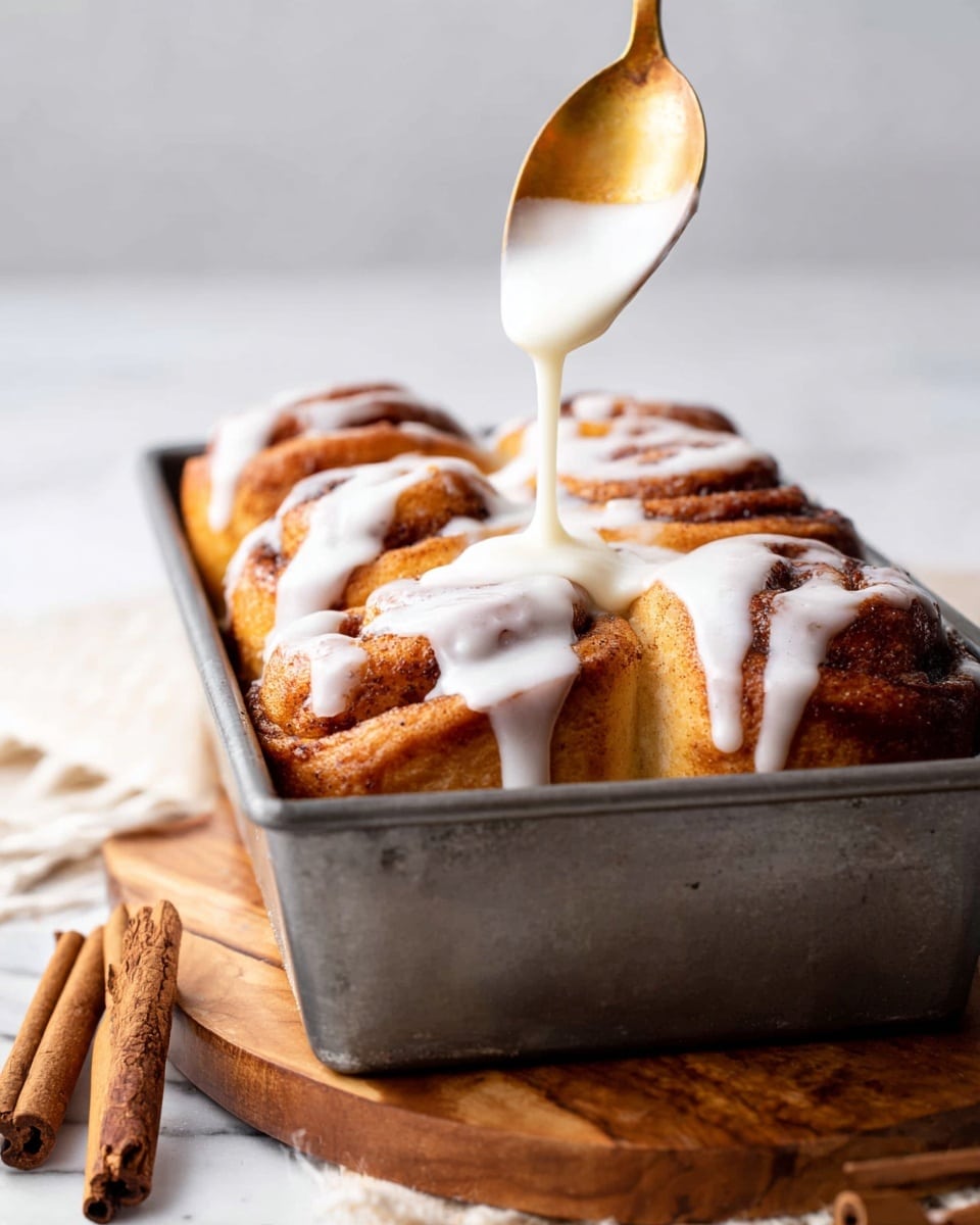The image shows a close-up view of a metal loaf pan filled with five golden brown cinnamon rolls standing upright next to each other. A golden spoon above pours a thick white glaze that drips down the tops of the cinnamon rolls in smooth streams. The cinnamon rolls have a rough, slightly crunchy texture with a darker, cinnamon-sprinkled outer layer. The pan sits on a wooden board with a cinnamon stick nearby, all set on a white marbled surface. Photo taken with an iphone --ar 4:5 --v 7