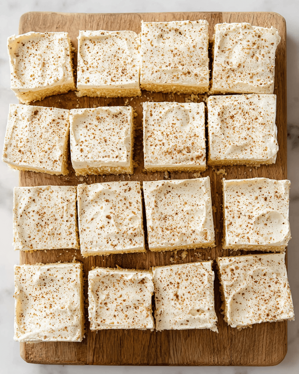 The image shows a rectangle wooden board on a white marbled surface, holding sixteen square pieces of cake arranged neatly in a 4 by 4 grid. Each piece has two layers: a light yellow cake base topped with a thick, creamy white frosting, which is sprinkled evenly with small brown specks that look like cinnamon or nutmeg. The texture of the frosting is smooth but with slight creamy ridges, while the cake appears soft and spongy with visible small air holes. The top edges of a few pieces are slightly uneven, adding a homemade feel. photo taken with an iphone --ar 4:5 --v 7
