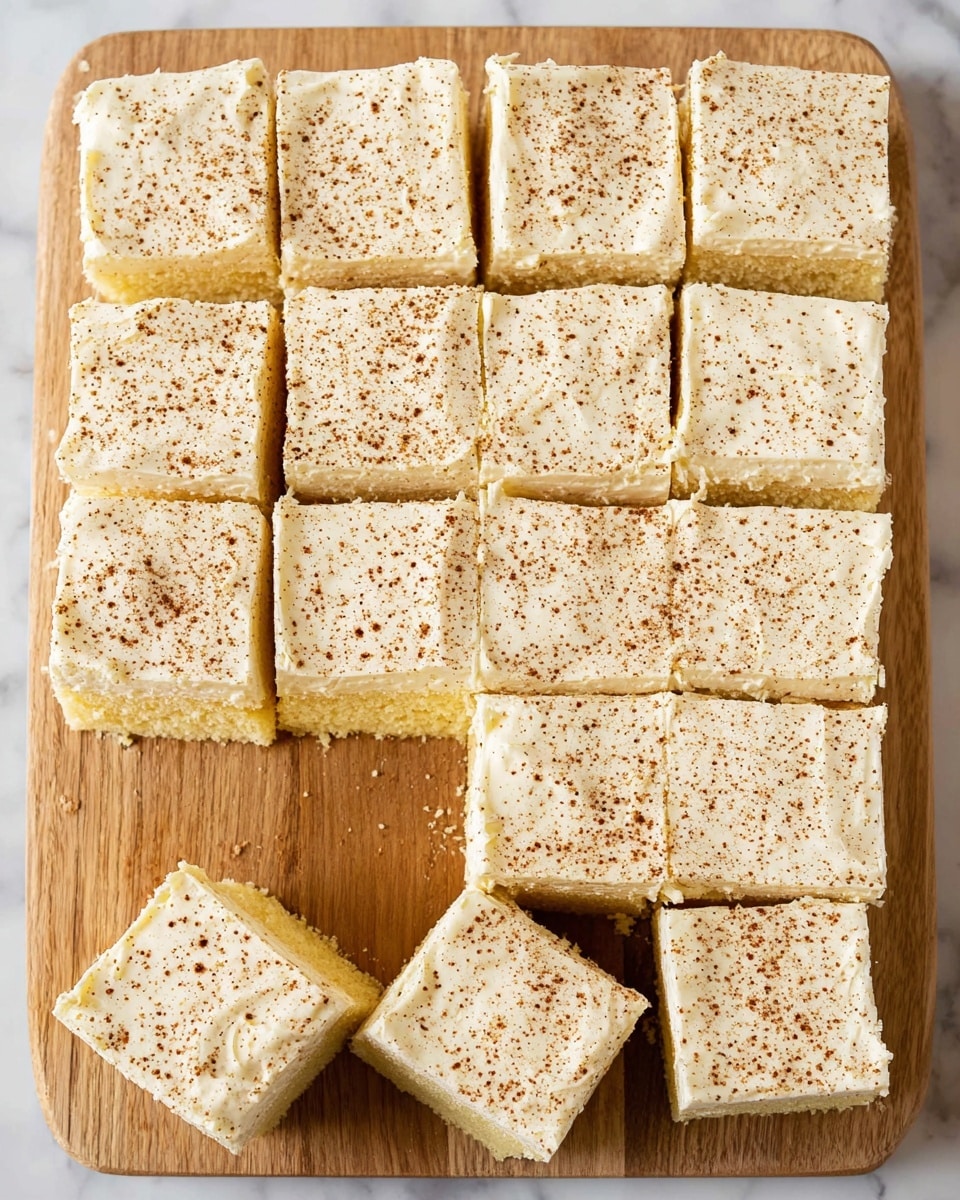 The image shows a wooden cutting board with sixteen square pieces of a light yellow cake with a creamy white frosting layer on top. Each piece is evenly cut, and the frosting is smooth but slightly textured with small air bubbles. The frosting is lightly sprinkled with brown spice powder evenly over all the pieces. The cake layers underneath the frosting have a soft, moist texture, and all squares are neatly placed close to each other on the board. The board rests on a white marbled surface. photo taken with an iphone --ar 4:5 --v 7