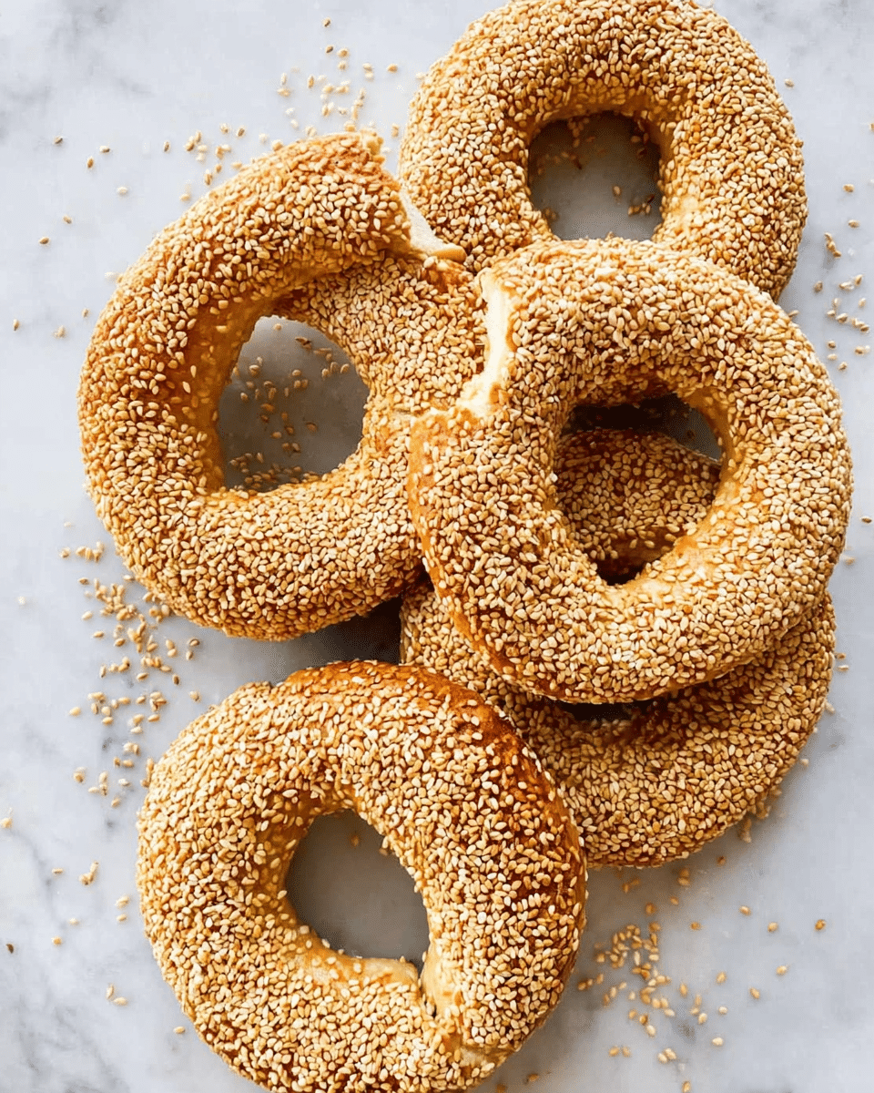 The image shows five round bagels covered fully with light tan sesame seeds on their crunchy surface. Each bagel has a golden brown crust beneath the seeds and a visible soft inner dough layer where a piece is broken off from one bagel. They lie close together on a white marbled textured surface with some scattered sesame seeds around them. The bagels look fresh with a glossy baked finish. Photo taken with an iphone --ar 4:5 --v 7