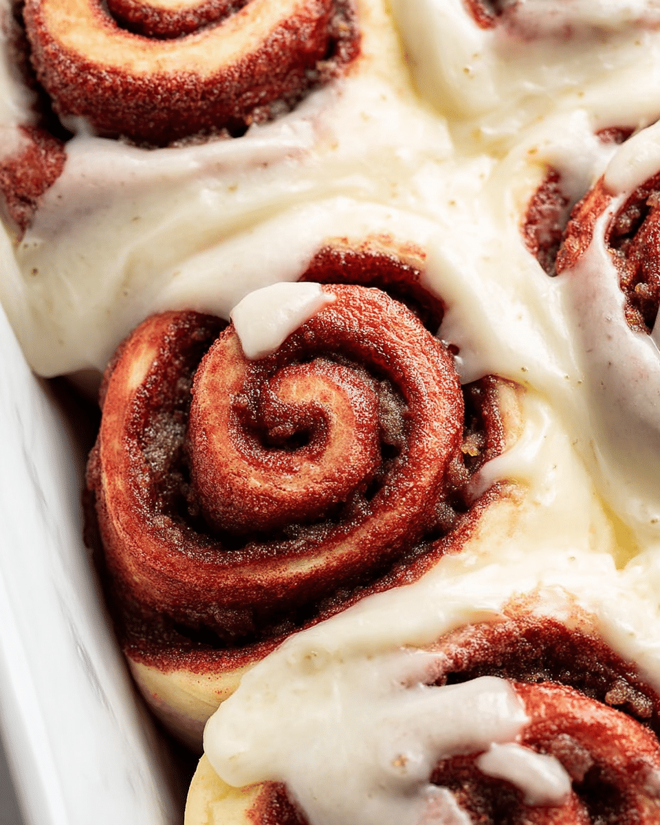 A close-up view of soft red velvet cinnamon rolls tightly packed in a white dish, each roll spiraled with dark brown cinnamon sugar filling, covered partially with a thick layer of creamy white icing that is smooth and slightly glossy, the icing pooling between and over parts of the rolls, some showing tiny vanilla bean specks, all set on a white marbled surface. photo taken with an iphone --ar 4:5 --v 7