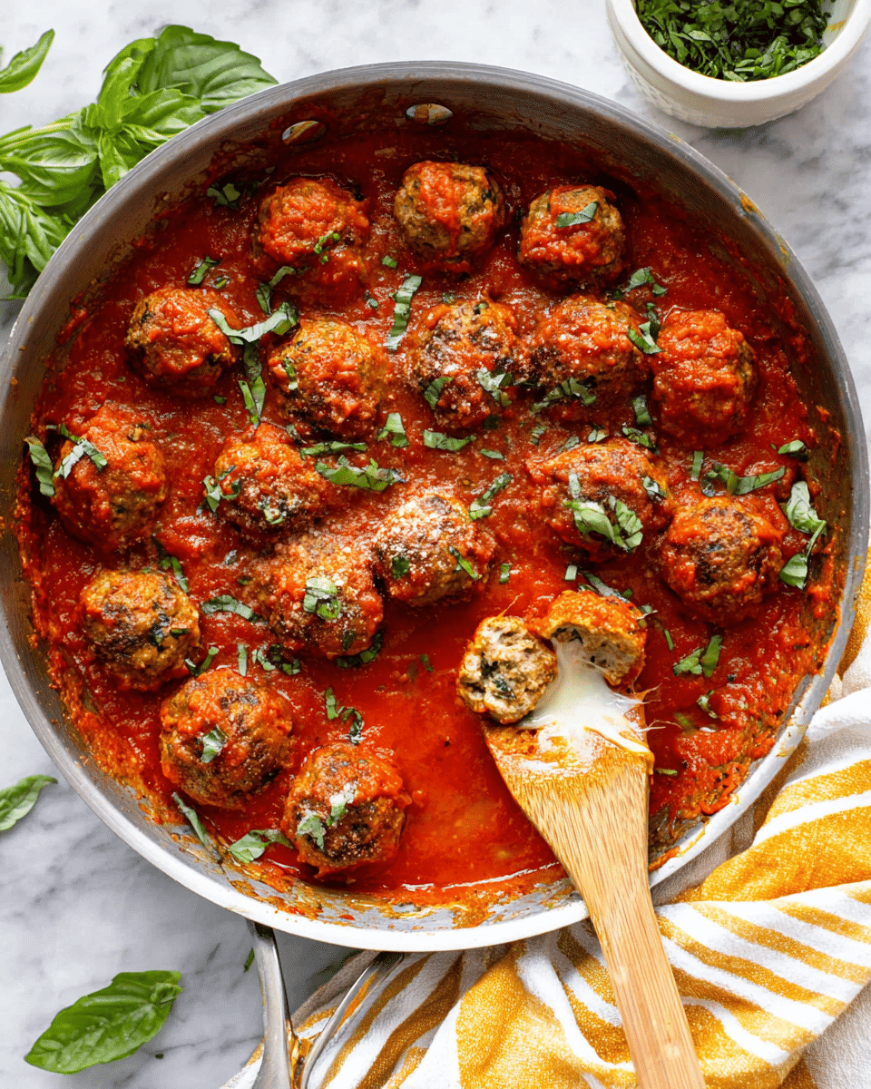 A silver pan filled with about fifteen round meatballs covered in thick, bright red tomato sauce, sprinkled with small green herb pieces, some whole green basil leaves on top, and one meatball cut open on a wooden spoon showing a gooey white cheese center, all placed on a white marbled surface with a small bowl of green herbs and a green leaf in the background and part of a striped cloth at the corner, photo taken with an iphone --ar 4:5 --v 7