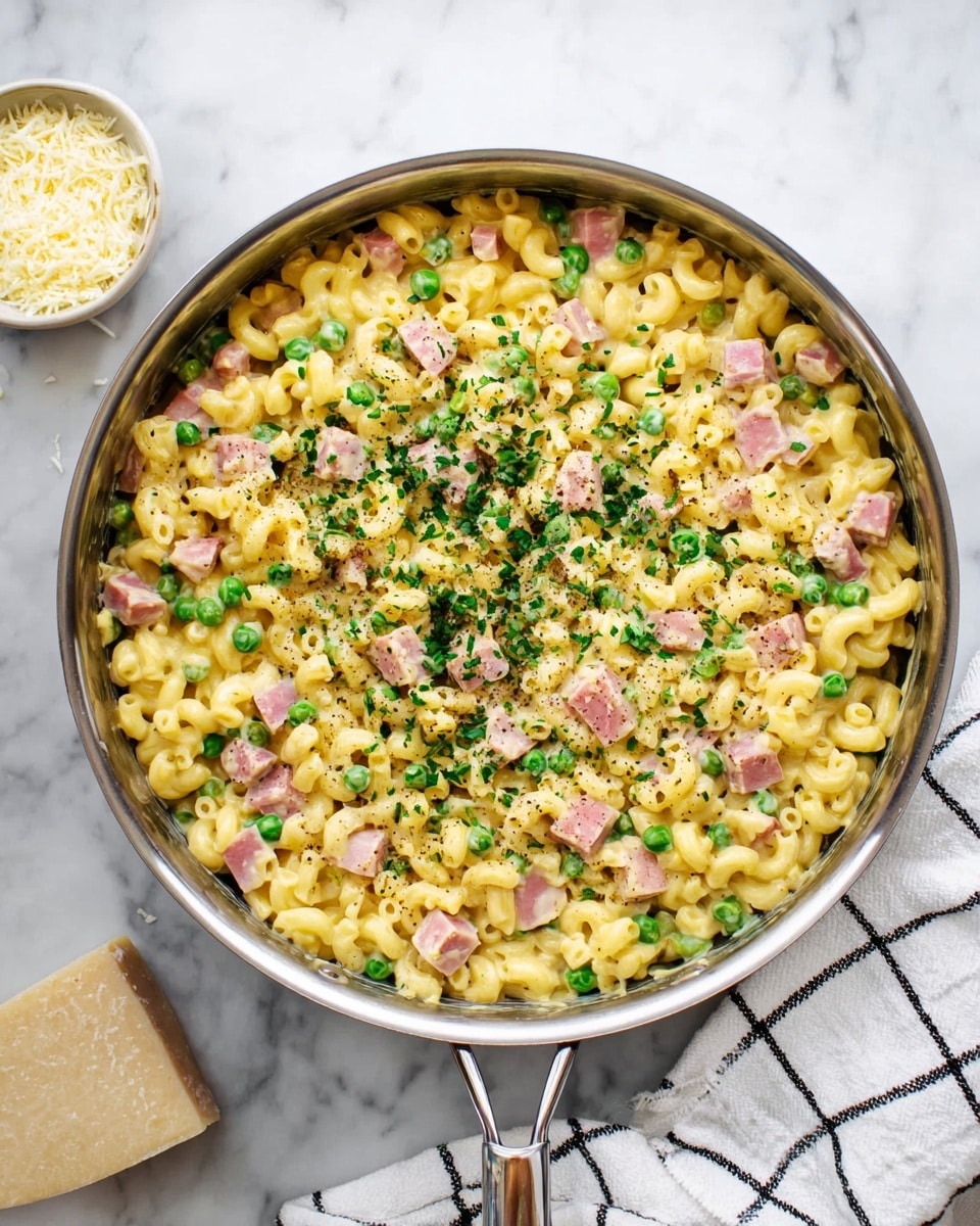 A large pan filled with a creamy macaroni dish that has three main visible layers: the base layer of yellow macaroni pasta, mixed evenly with light green peas and pink cubes of cooked ham; the second layer has sprinkled bright green chopped herbs across the top, adding color contrast; the final layer is a light dusting of grated pale yellow cheese and black pepper. The pan sits on a white marbled surface with a white cloth featuring a black grid pattern on the right side and a small bowl of grated cheese on the left. The photo is bright, showing the glossy texture of the pasta and the fresh look of the ingredients. Photo taken with an iphone --ar 4:5 --v 7