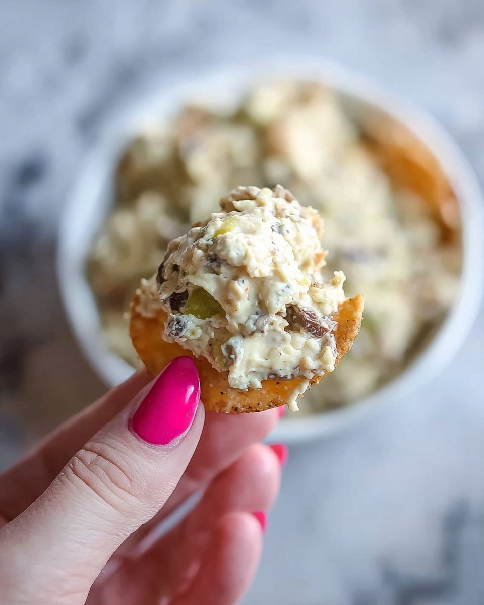 A close-up image of a pale golden, round chip held by a woman's hand with bright pink nail polish, topped with a creamy, chunky mixture that includes light beige and off-white ingredients with small bits of green and dark brown throughout. Below, a white bowl filled with the same creamy mixture sits on a white marbled surface, slightly out of focus in the background. The creamy topping looks thick and textured, with visible pieces of what might be vegetables or nuts. Photo taken with an iphone --ar 4:5 --v 7