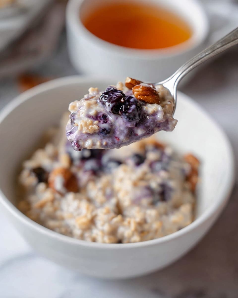 A close-up of a silver spoon holding a mix of two-layered oatmeal, the bottom layer is creamy white and the top layer is a light purple with visible dark berries and bits of nuts, hovering over a white bowl filled with the same oatmeal mixture on a white marbled surface, with a blurred white bowl of amber-colored liquid in the background. photo taken with an iphone --ar 4:5 --v 7