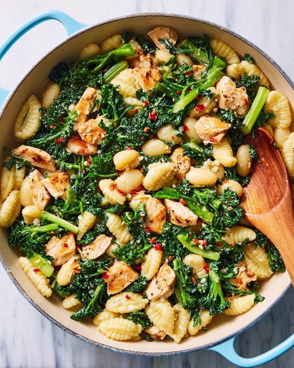 A light blue cast iron pan filled with three main layers: the first layer is pale yellow gnocchetti pasta shaped like small ridged tubes, spread evenly across the pan; the second layer consists of bright green broccoli rabe and kale pieces, mixed throughout and adding texture with leafy and stem parts; the third layer has golden-brown cooked chicken chunks scattered on top, some sprinkled with small bits of red chili flakes. A wooden spoon is resting inside the pan on the right side. The whole pan is on a white marbled surface. photo taken with an iphone --ar 4:5 --v 7