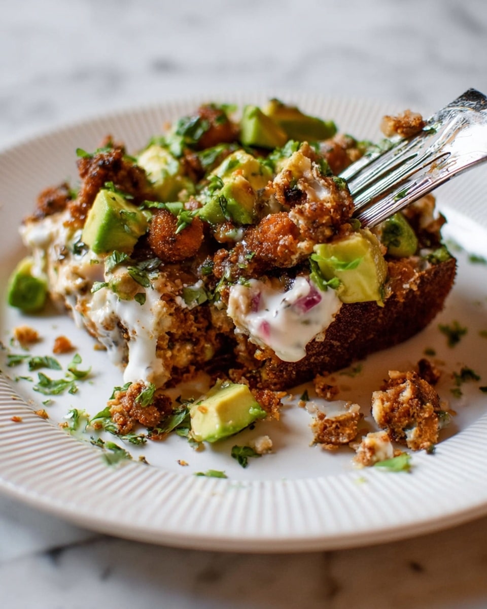 The dish shows two layers, with the bottom layer being a dark brown base that looks like toasted bread or a cake and the top layer made of chunky brown pieces that might be cooked meat or textured protein. Scattered across the top are bright green cubes of avocado and some chopped fresh green herbs. A white creamy sauce is drizzled over the top and pools slightly on the white, ridged plate, giving a soft contrast to the crispy texture above. A fork is partially pressing into the dish, picking up a mix of the mashed creamy part and chunks from the layers. The plate sits on a white marbled surface with small crumbs and flecks of herbs around it. photo taken with an iphone --ar 4:5 --v 7