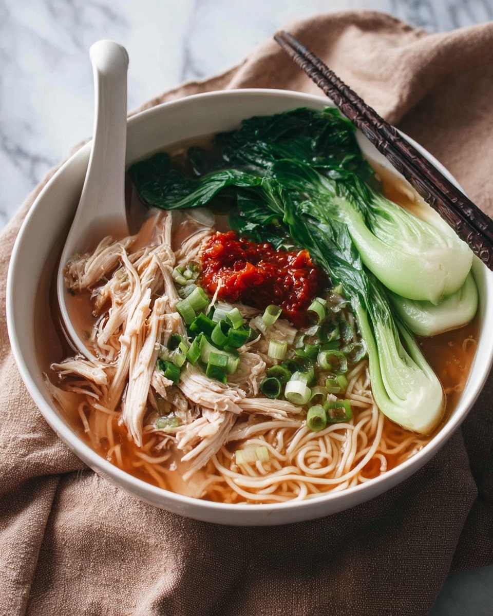 A white bowl filled with clear brown broth at the base, topped with light beige noodles that fill most of the bowl. On one side, there are tender white shredded chicken pieces arranged over the noodles. Above the chicken, bright red chili paste adds a splash of color. Fresh green chopped scallions are sprinkled on top. On one edge, large dark green leafy bok choy rests partially submerged in the broth. A white ceramic soup spoon is placed inside the bowl on the left side, and a pair of dark chopsticks is placed on the right edge. The bowl sits on a soft brown cloth with a white marbled surface behind it. Photo taken with an iphone --ar 4:5 --v 7