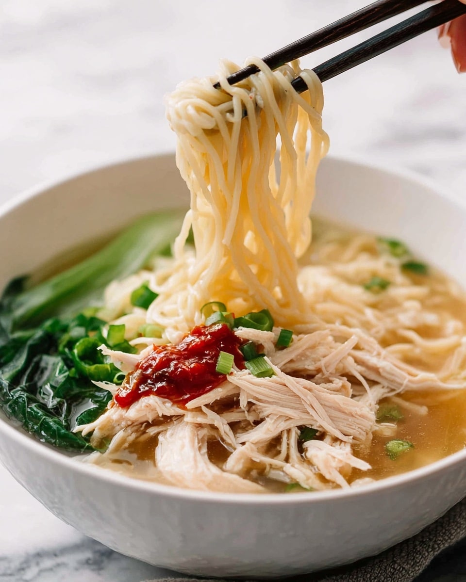 A white bowl filled with clear broth and thin, pale yellow noodles sitting in the soup as the main layer. On top of the noodles are shredded pieces of white chicken meat scattered around. Bright green chopped spring onions and leafy greens add color on one side. A dollop of red chili sauce rests near the chicken for a spicy touch. Black chopsticks held by a woman's hand lift a small bundle of noodles in the air above the bowl. The background has a soft white marbled texture, giving a clean, fresh feel. photo taken with an iphone --ar 4:5 --v 7
