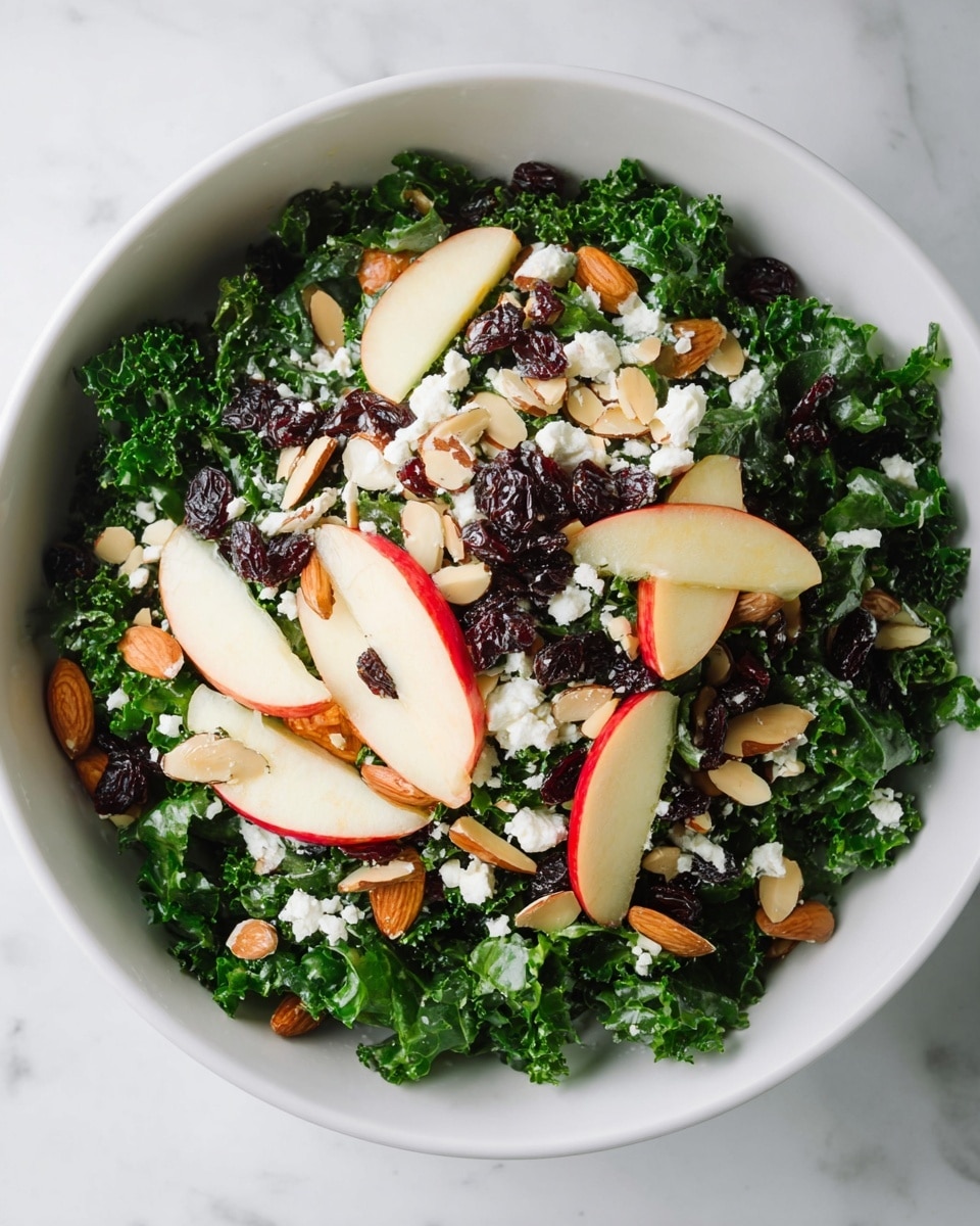 A white round bowl filled with a fresh kale salad showing three main layers: the bottom layer is dark green kale leaves with a curly texture, the middle layer has thin red and white apple slices evenly placed, and the top layer is sprinkled with small white crumbles of cheese, glossy dark raisins, and light brown sliced almonds scattered all around. The bowl sits on a white marbled surface. photo taken with an iphone --ar 4:5 --v 7